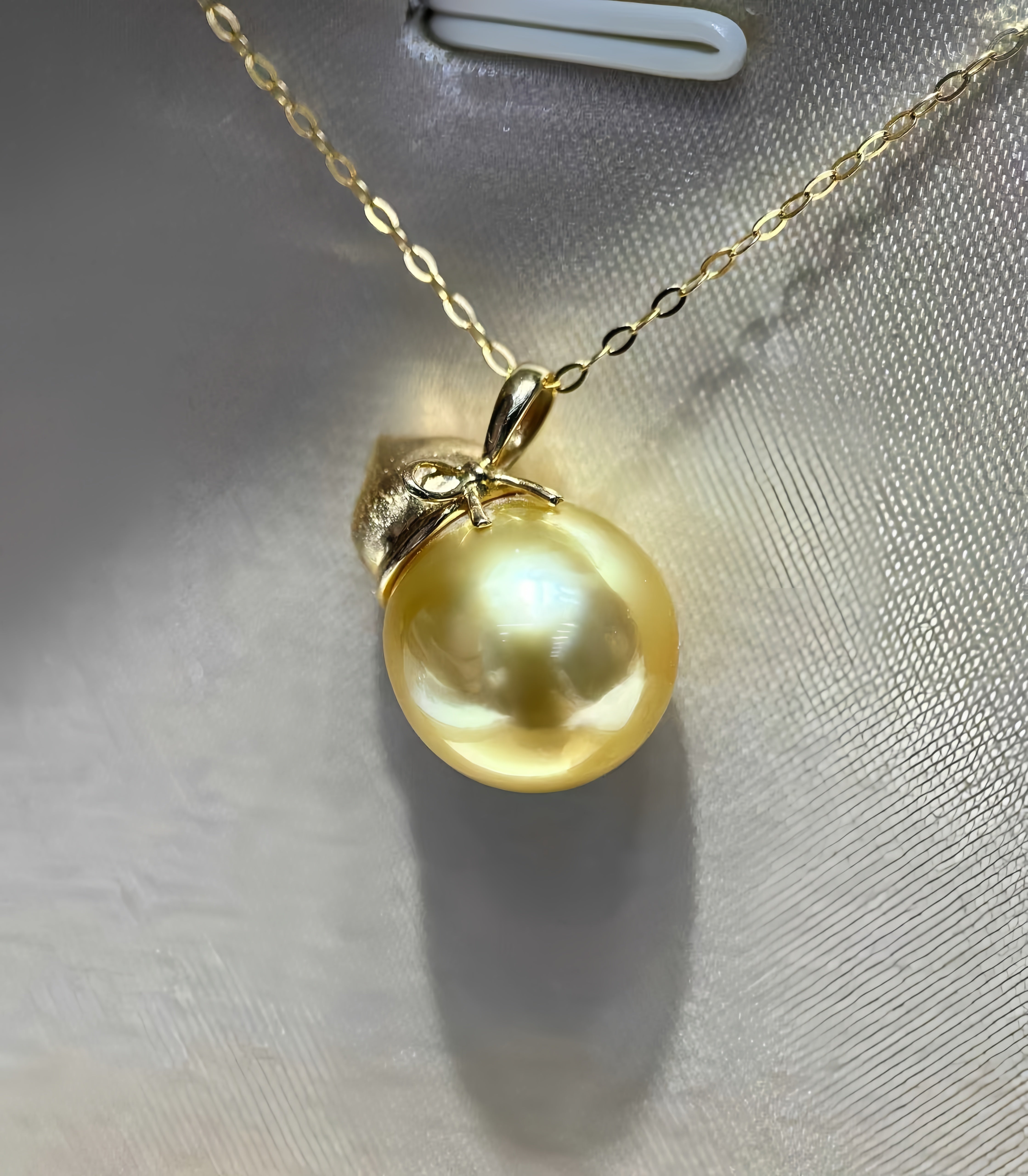 A gold chain necklace featuring a large, lustrous yellow pearl pendant with a gold cap, displayed on a textured light gray surface.