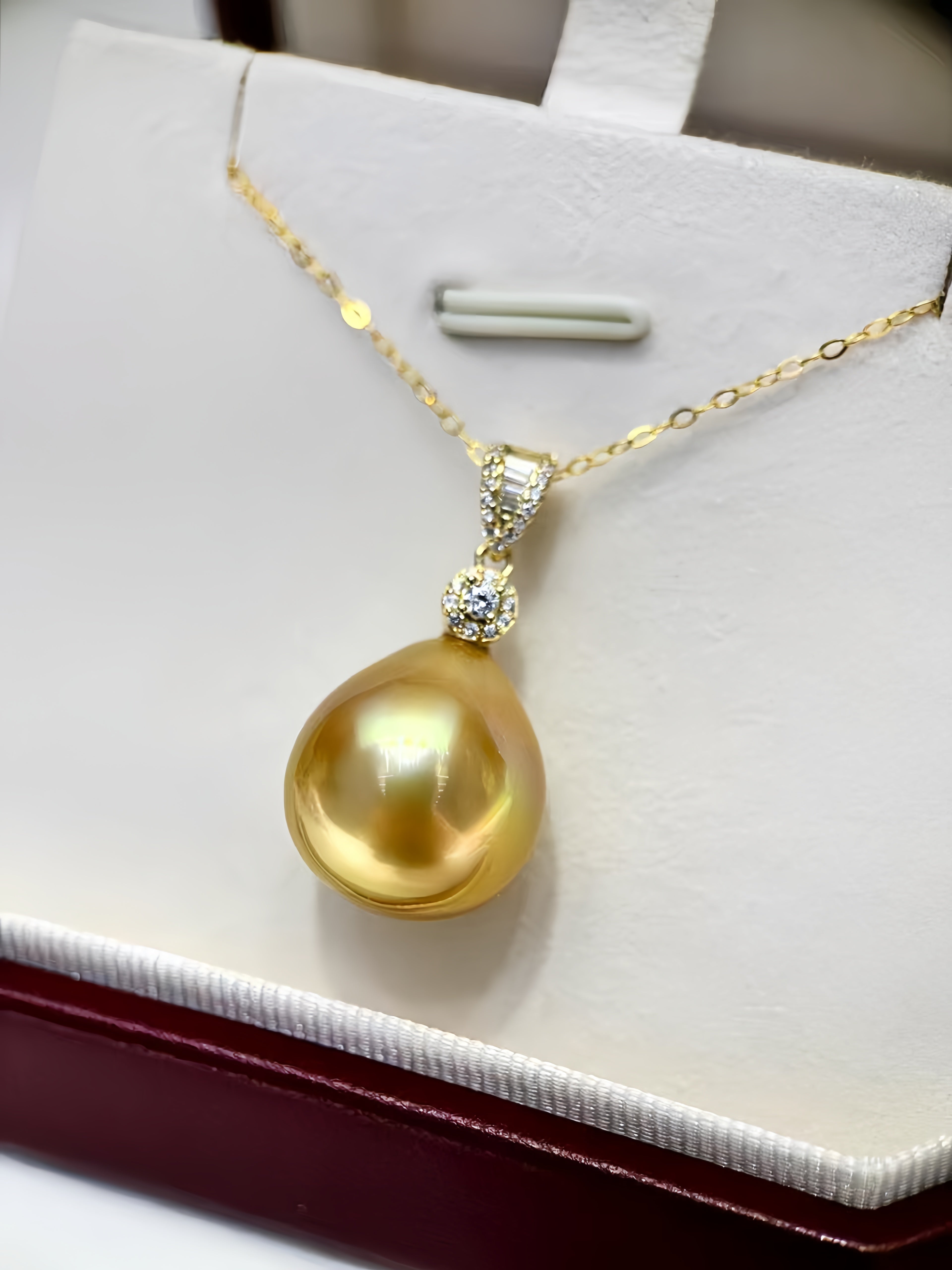A gold pendant necklace featuring a large, lustrous yellow pearl pendant and a small halo of clear stones, displayed on a white jewelry box.