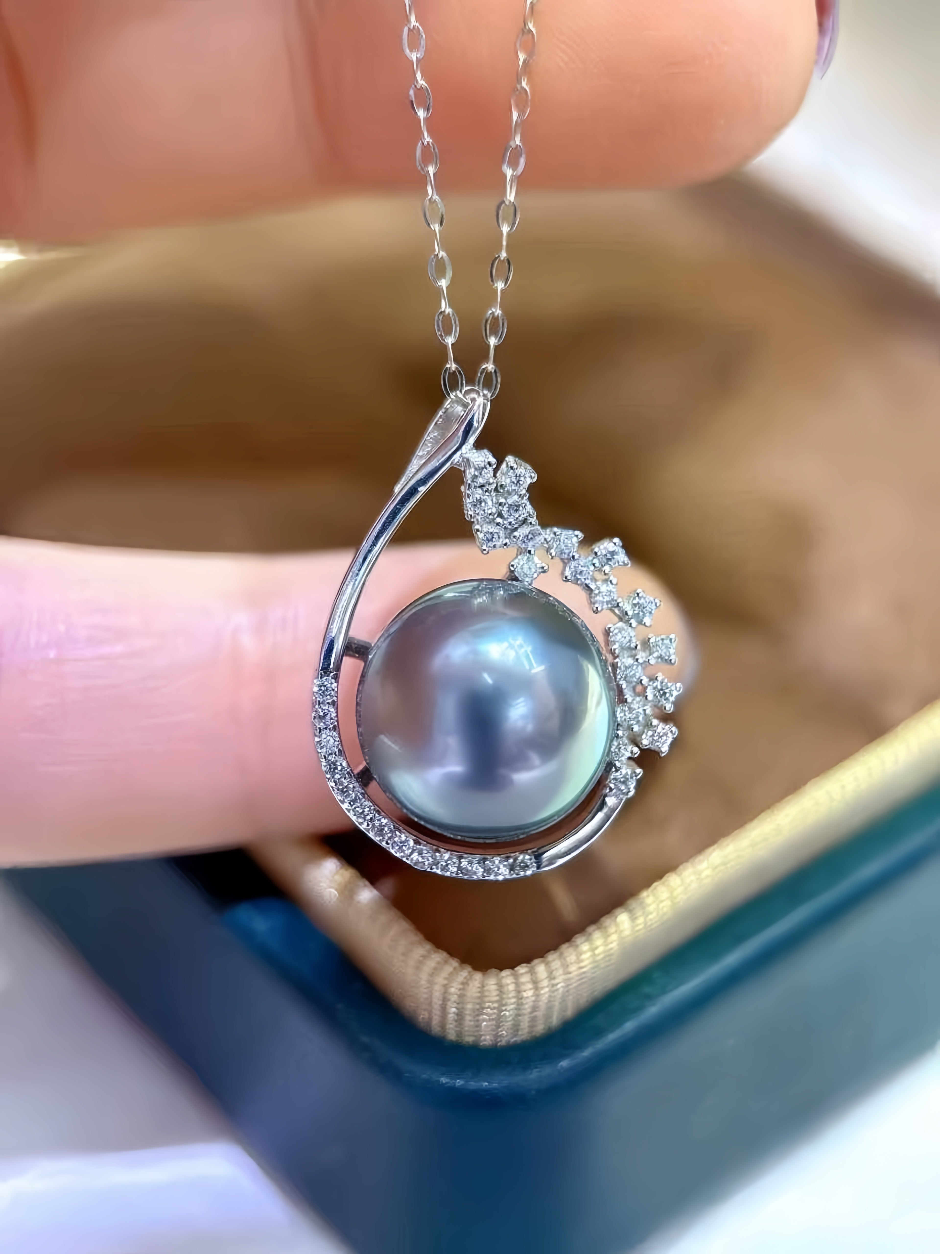 A close-up of a silver pendant necklace featuring a large round gray pearl and small clear stones, held between fingers above an open jewelry box.