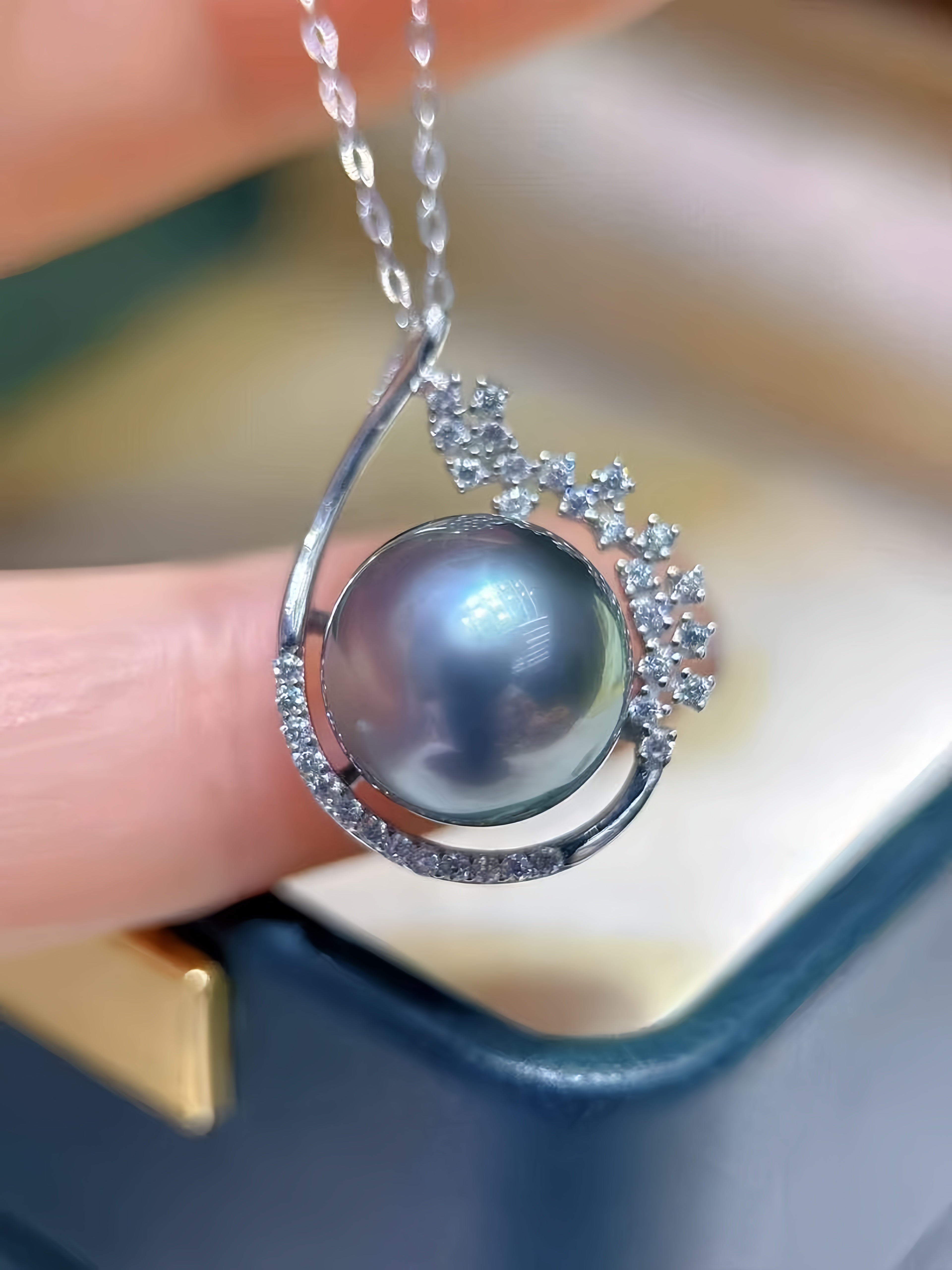A close-up of a silver pendant necklace featuring a large round gray pearl, surrounded by a crescent-shaped silver frame set with small clear stones, held by a finger above a jewelry box.
