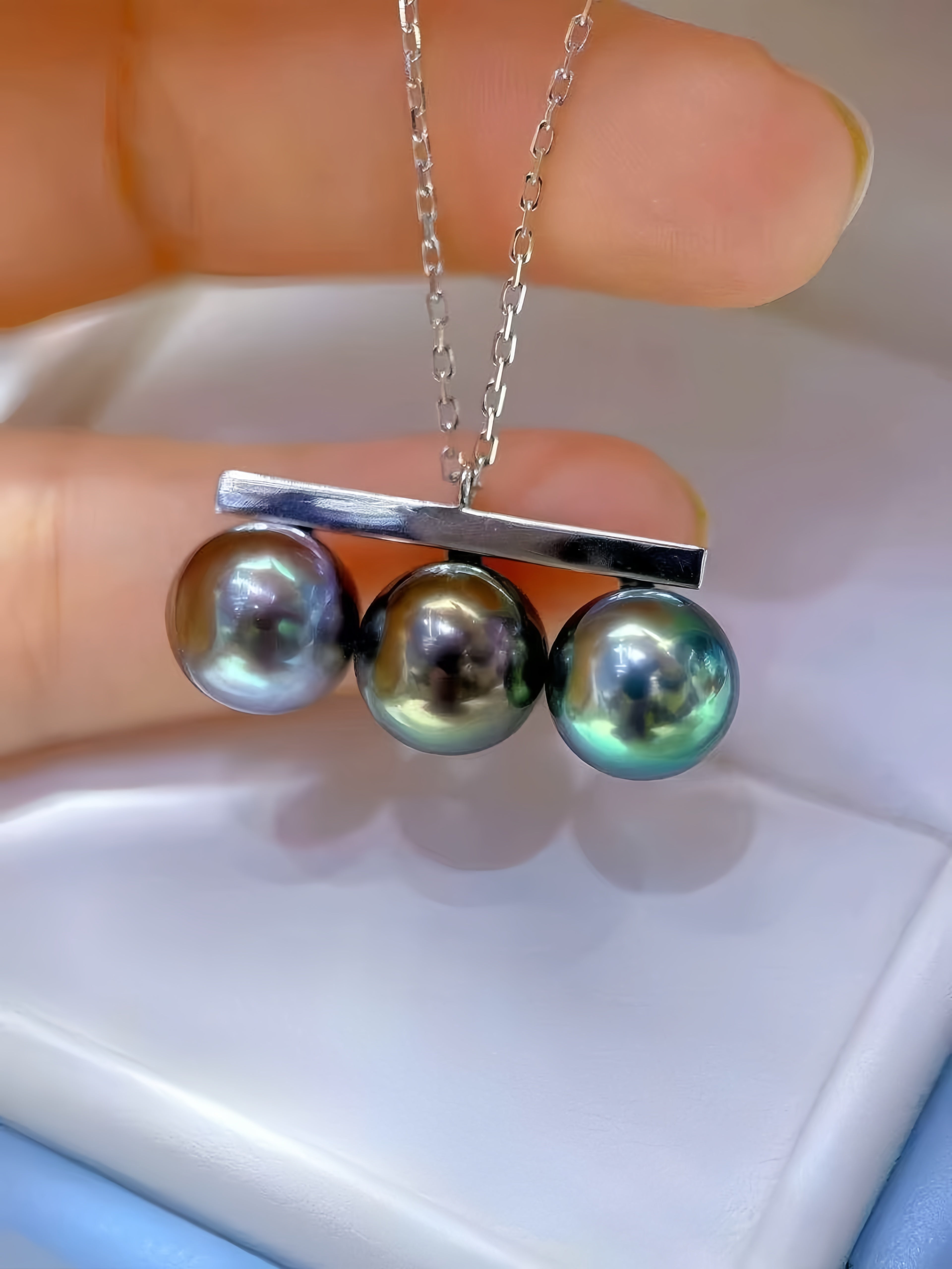 A close-up of a silver necklace with three round iridescent freshwater pearls hanging from a horizontal bar, held between fingers over a white jewelry box.