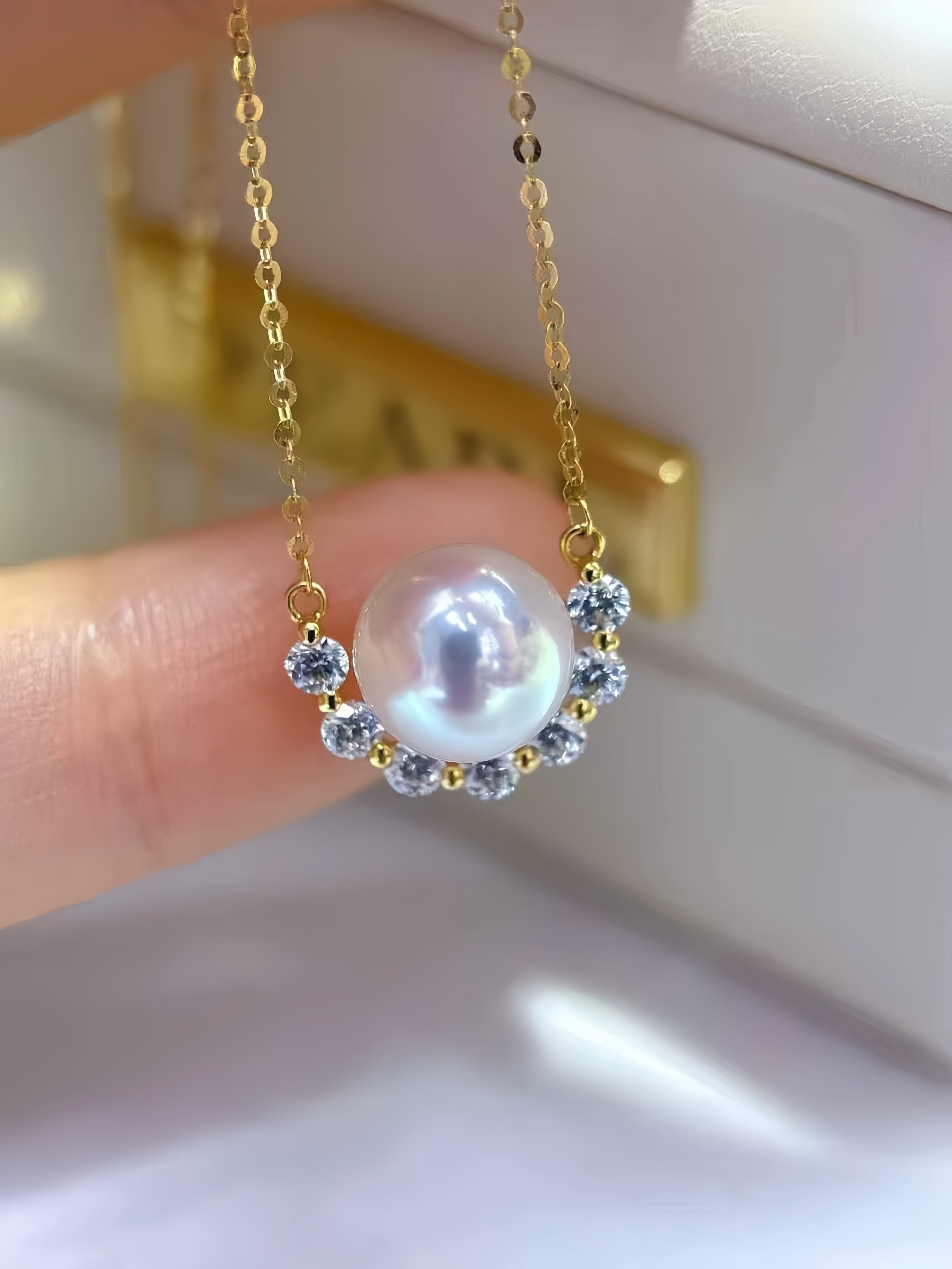 A gold chain necklace with a large white pearl pendant, accented by small clear stones in a crescent shape, held by a finger against a soft-focus jewelry box background.