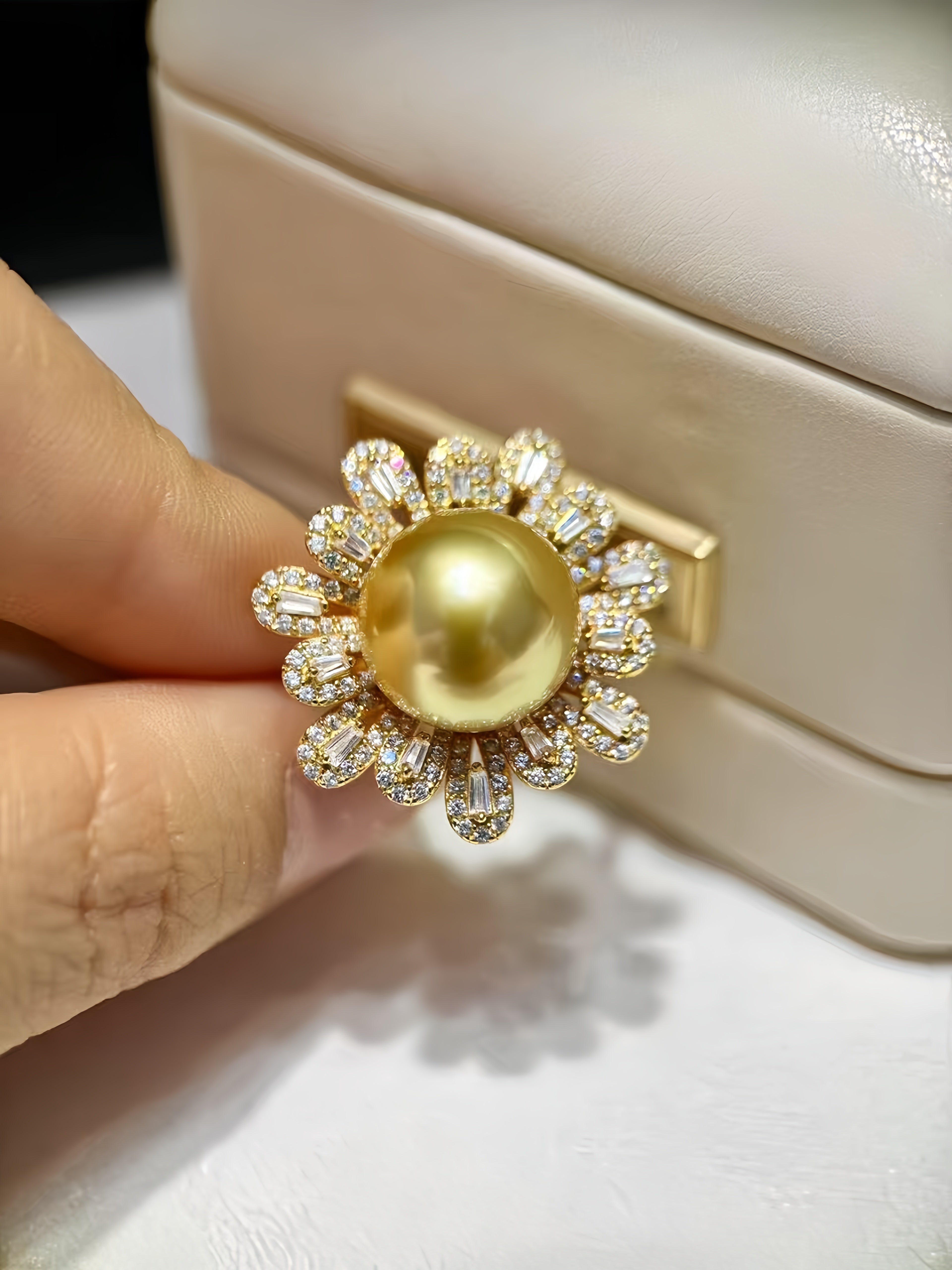 A gold ring featuring a large round yellow pearl in the center, surrounded by a halo of small clear baguette and round-cut stones, held by a hand in front of a cream-colored jewelry box.