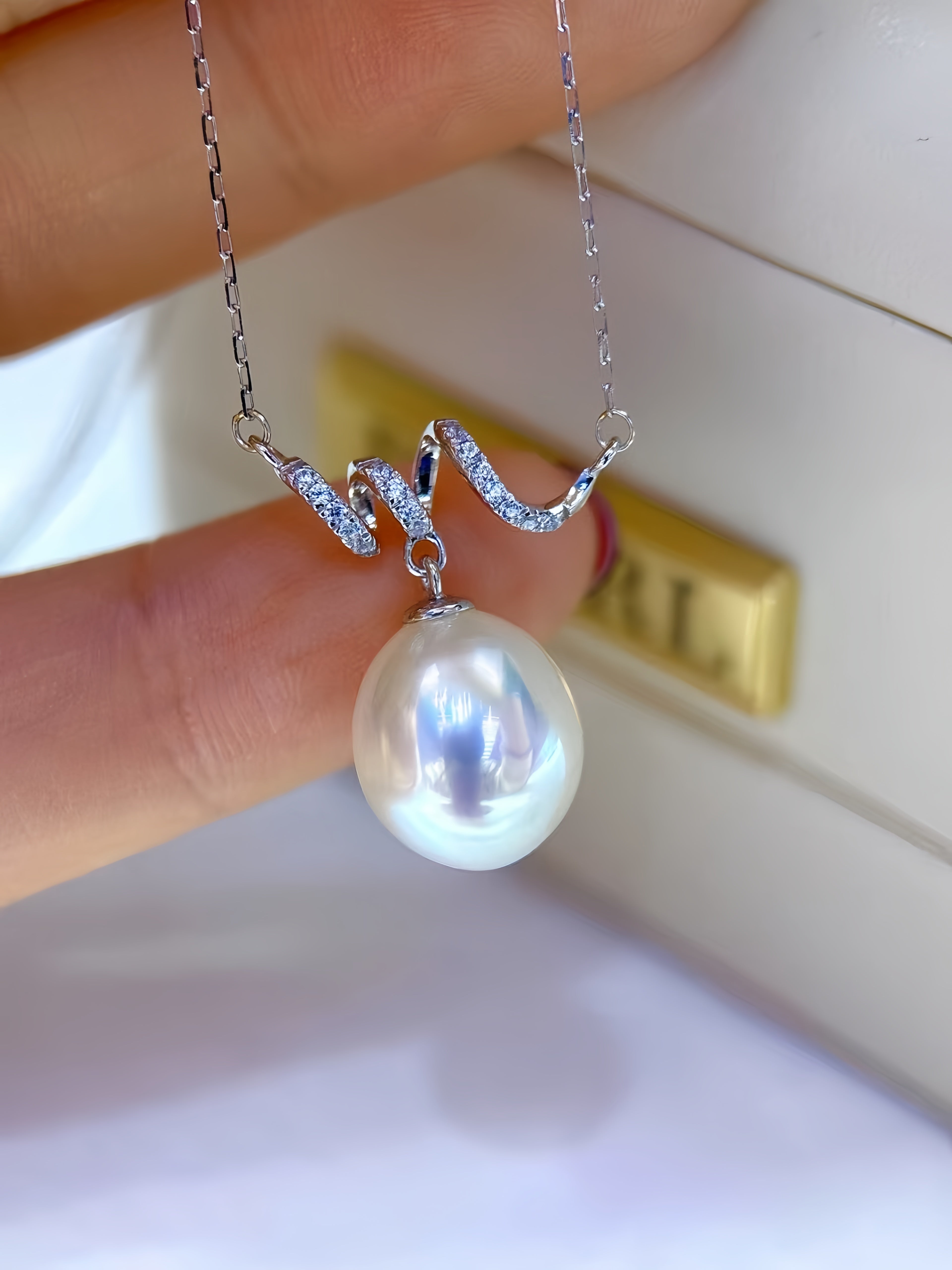 A close-up of a silver chain necklace with a large, lustrous white freshwater pearl pendant and a wavy silver top featuring small clear stones, held between fingers.