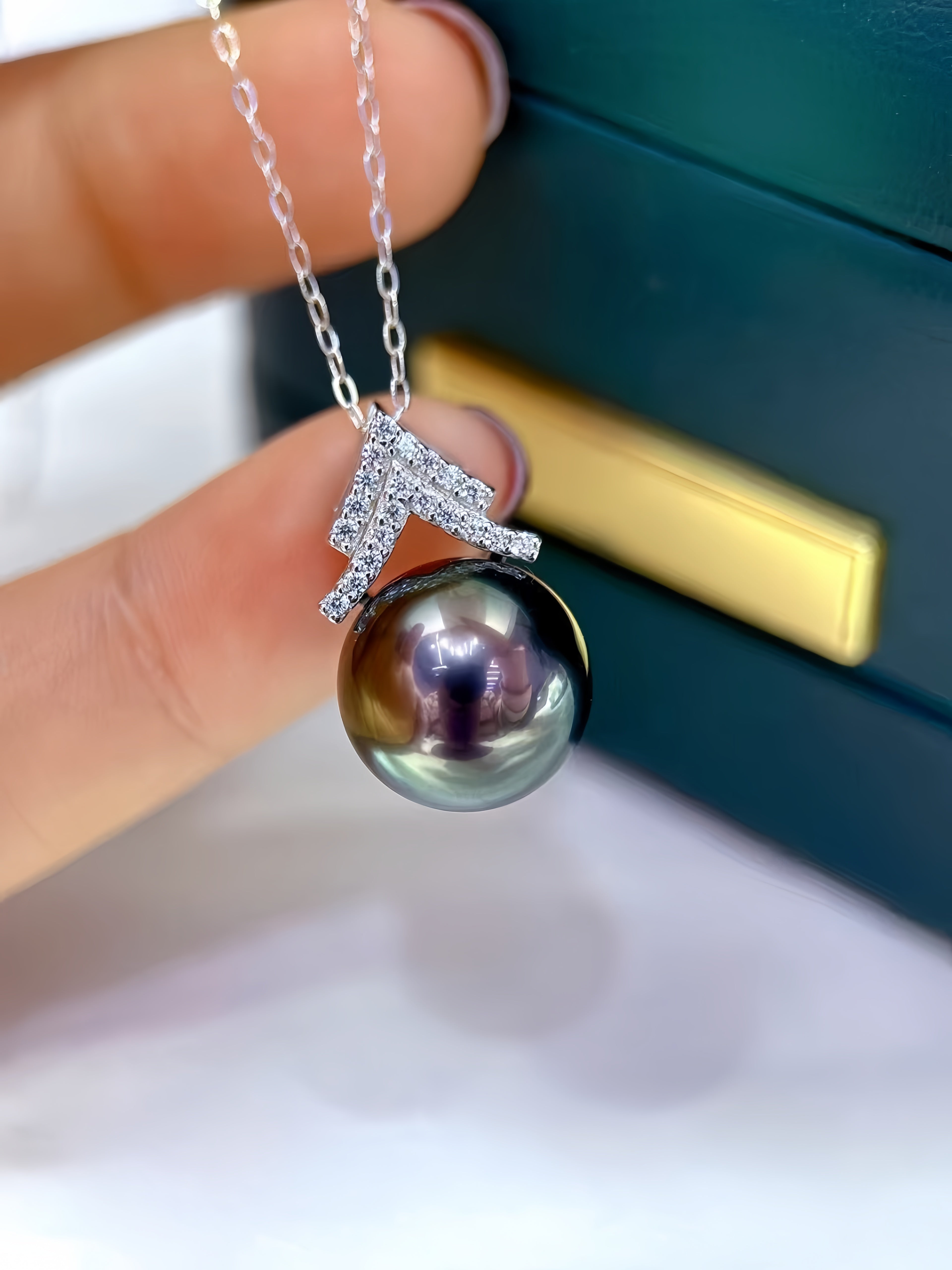 A close-up of a hand holding a silver pendant necklace with a large round dark pearl and a sparkling geometric silver top, with a dark teal jewelry box in the background.