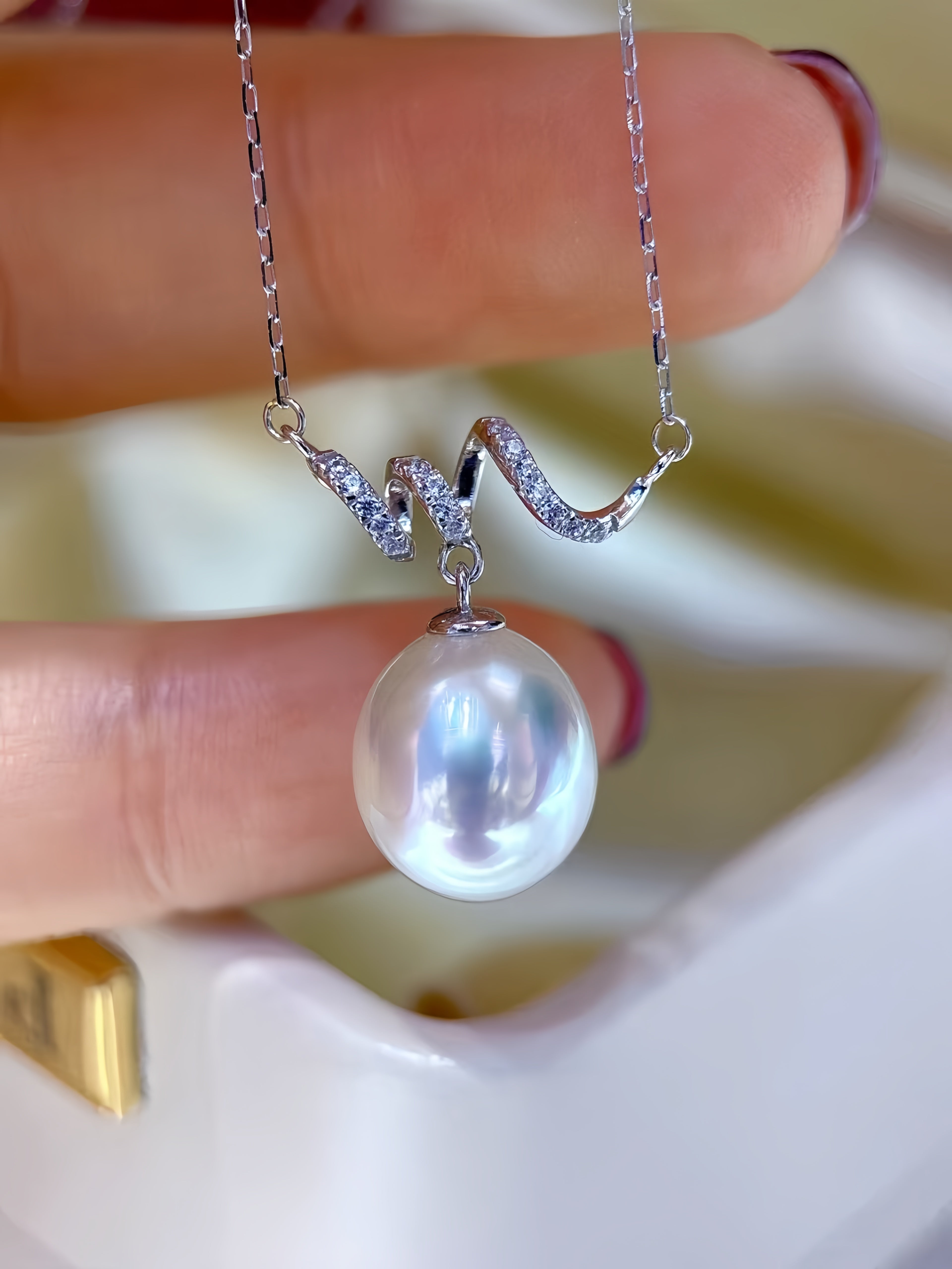 A close-up of a silver chain necklace with a wavy pendant featuring small clear stones and a large lustrous pearl pendant, held between fingers.