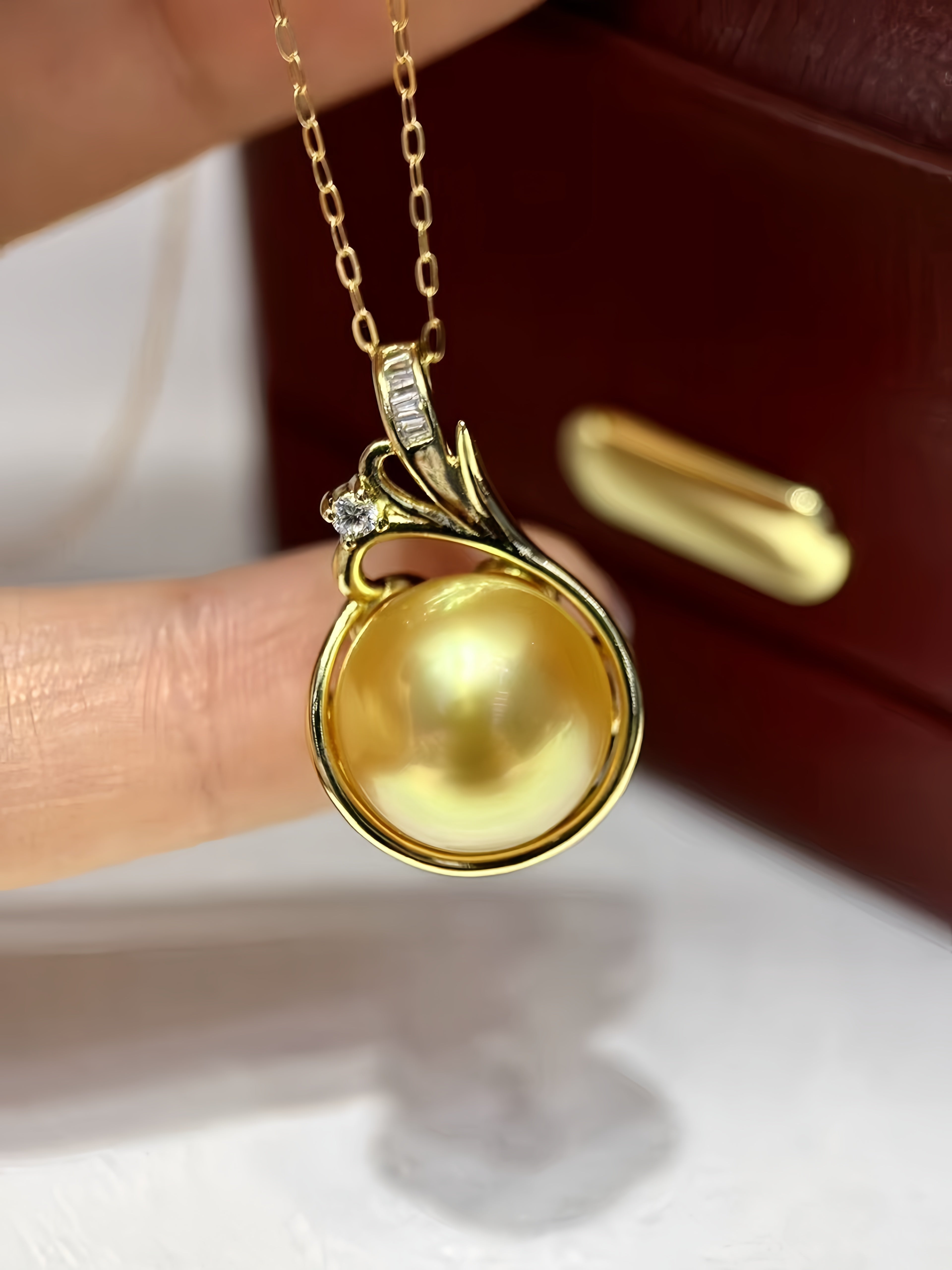 A gold pendant necklace featuring a large, round, lustrous yellow pearl, held by fingers in front of a dark brown leather jewelry box.
