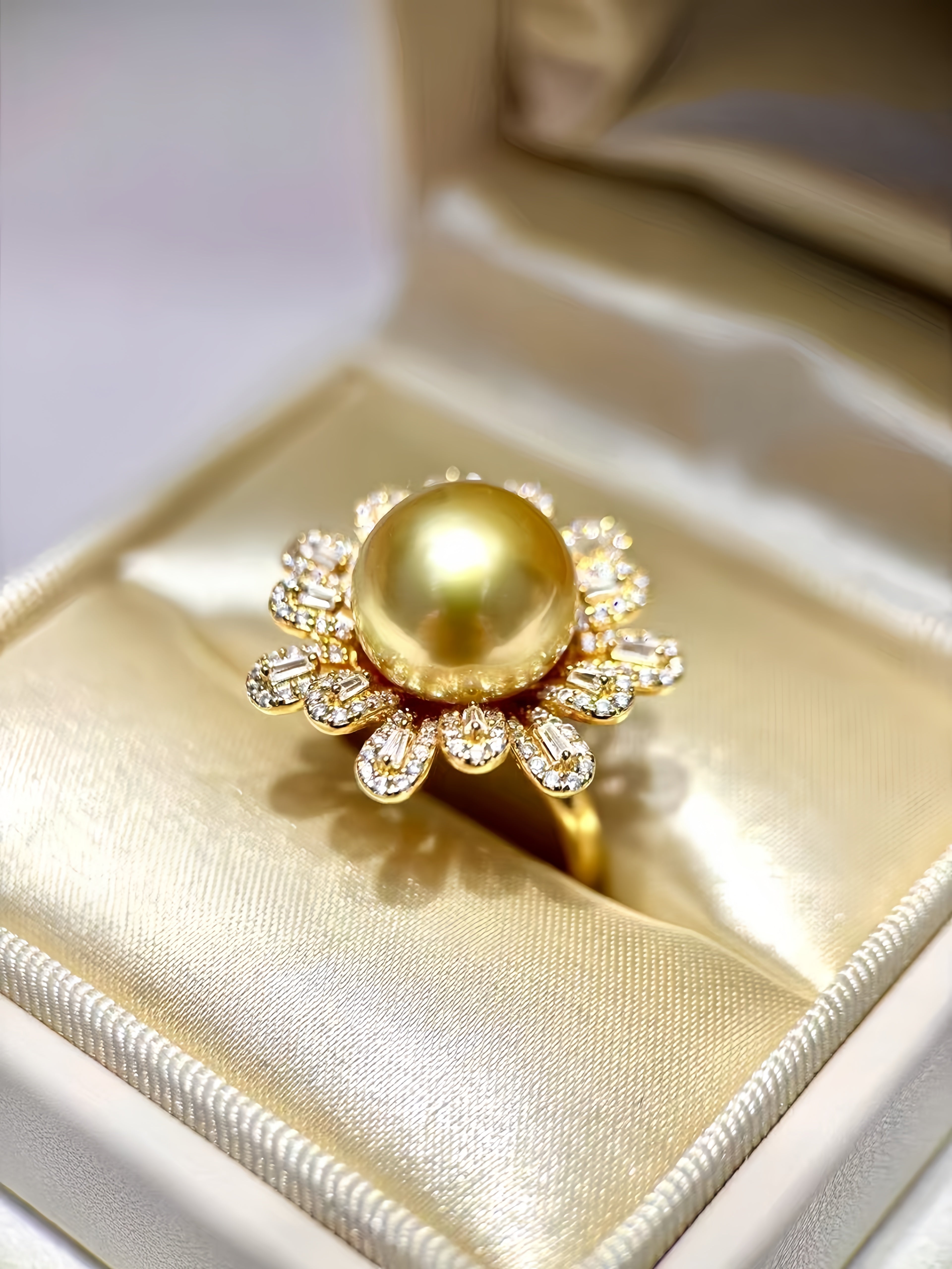 A gold ring featuring a large round pearl surrounded by small clear stones, displayed in an open cream satin ring box.