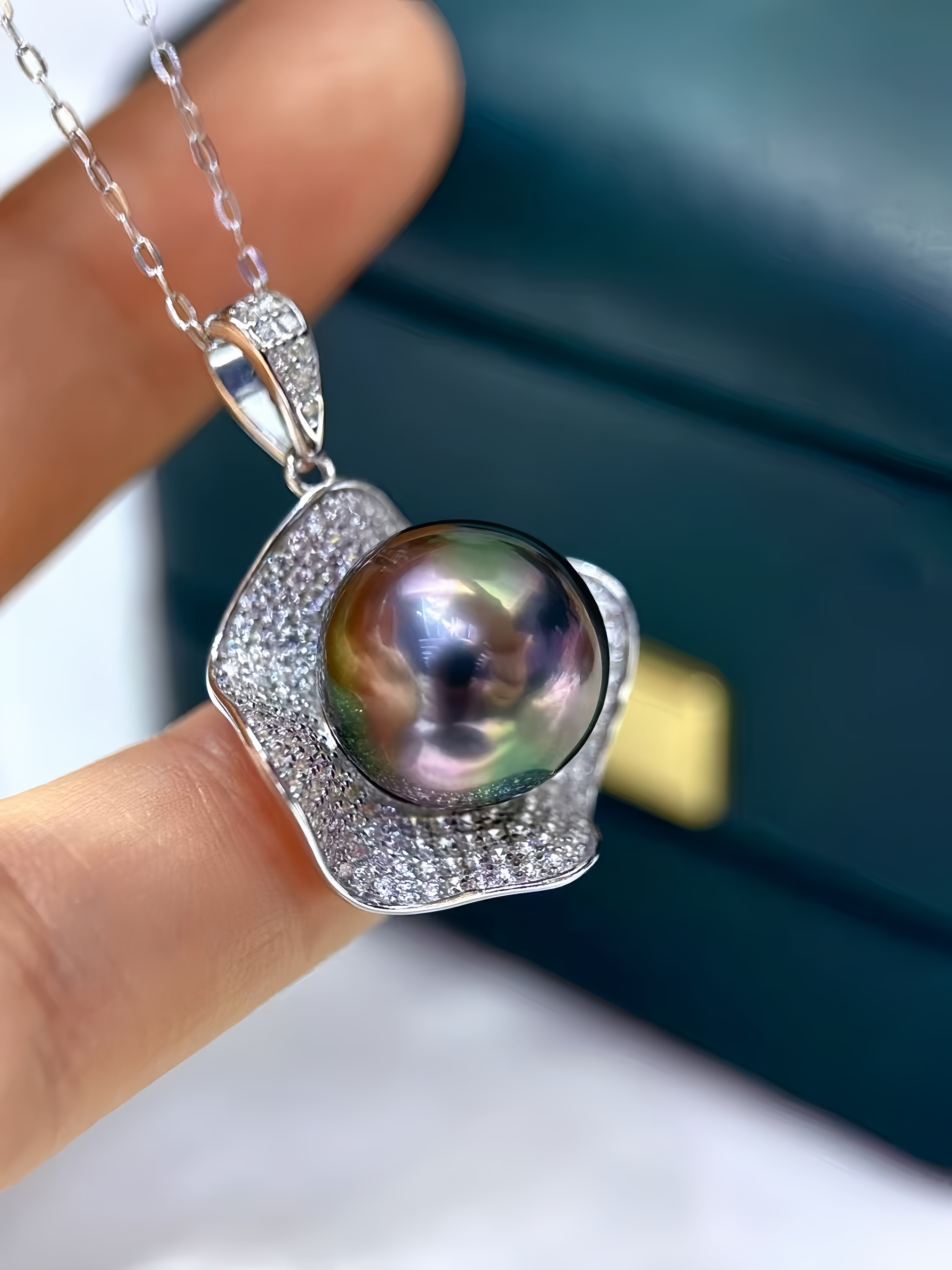 A close-up of a hand holding a silver pendant necklace featuring a large, iridescent pearl surrounded by small clear stones, with a dark teal jewelry box blurred in the background.