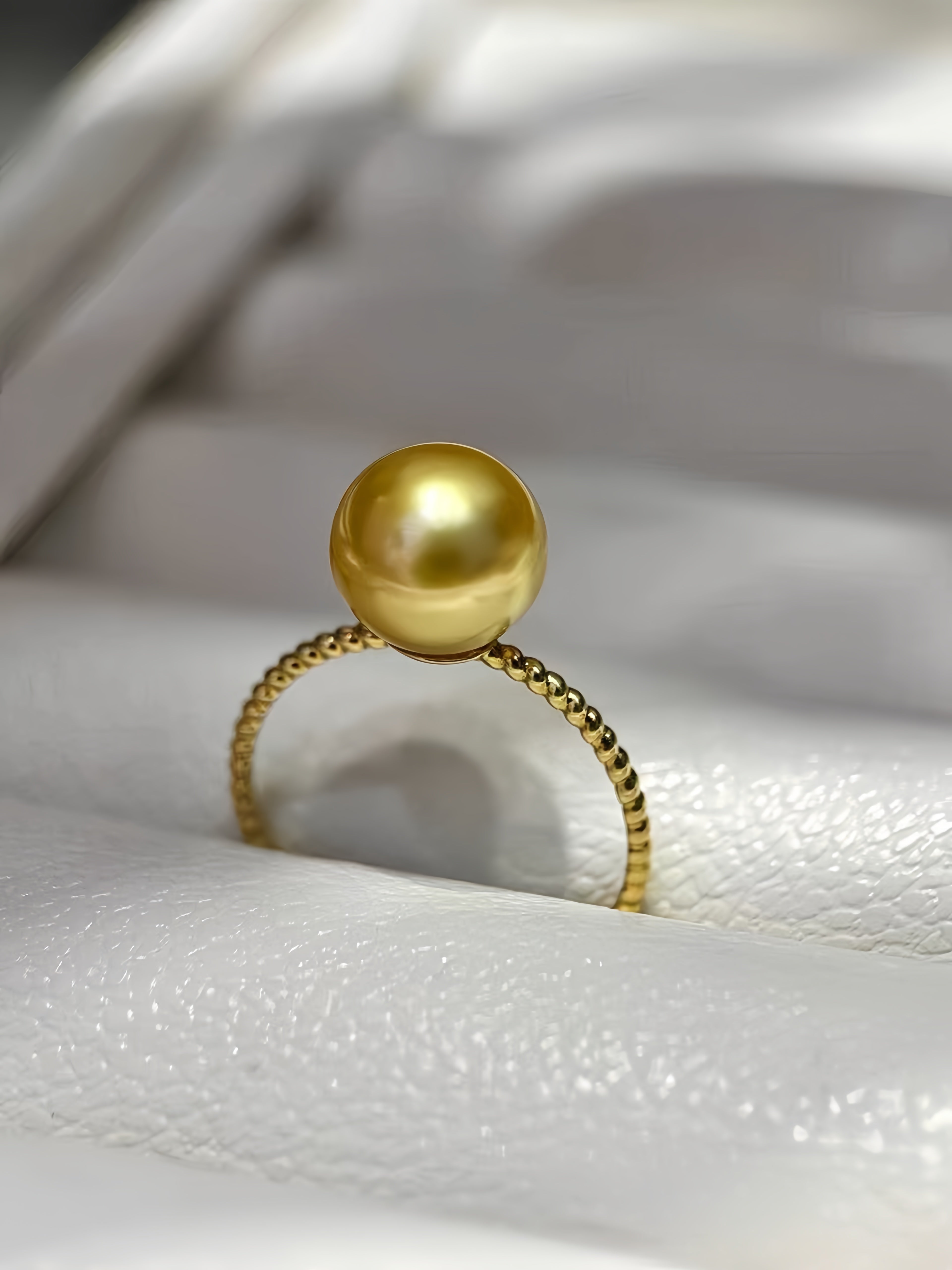 A gold ring featuring a large, round, lustrous gold pearl on a textured gold band, displayed on a white textured surface.