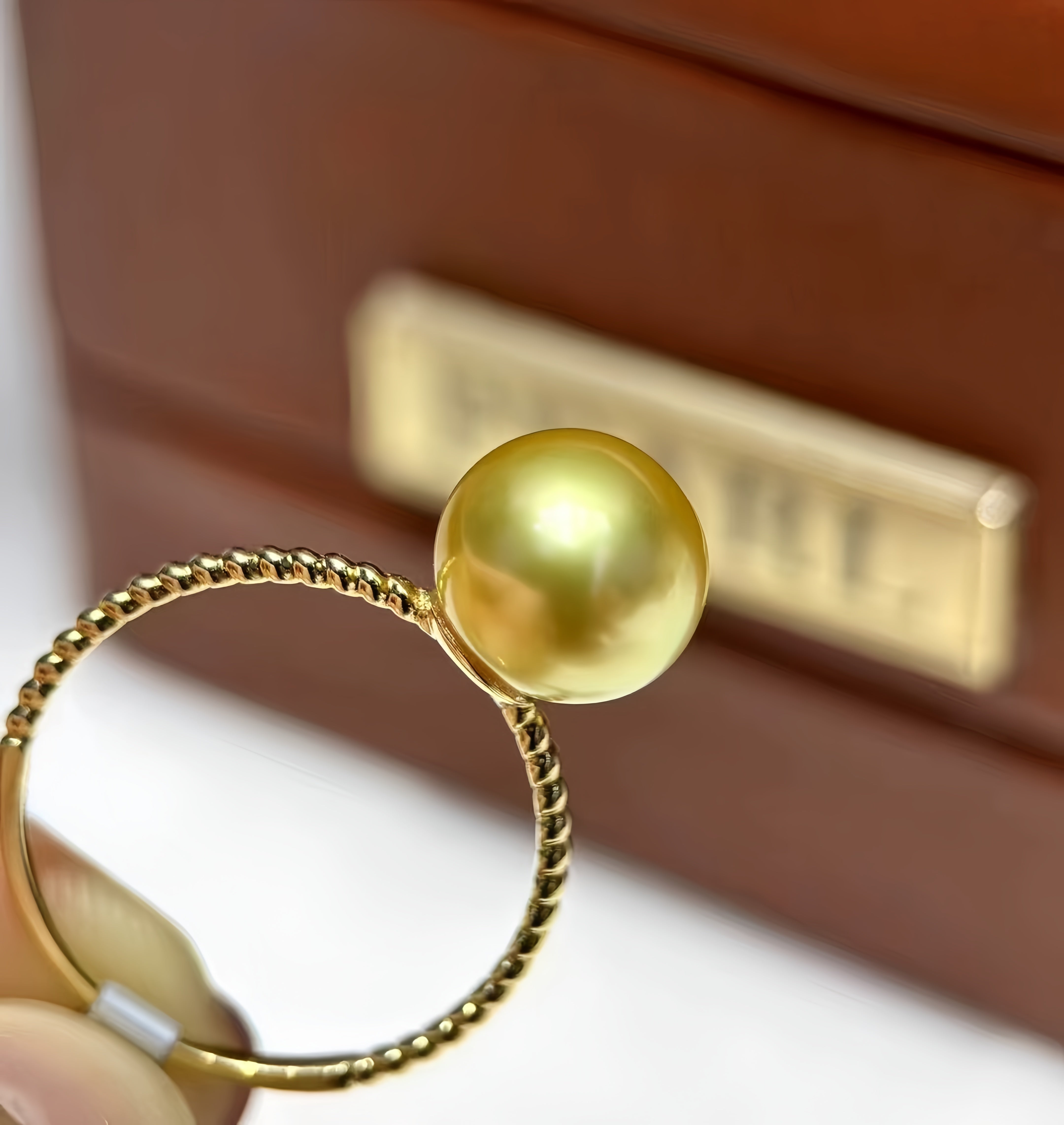 A gold ring with a large, round, lustrous yellow gold pearl, held by a hand in front of a brown jewelry box.