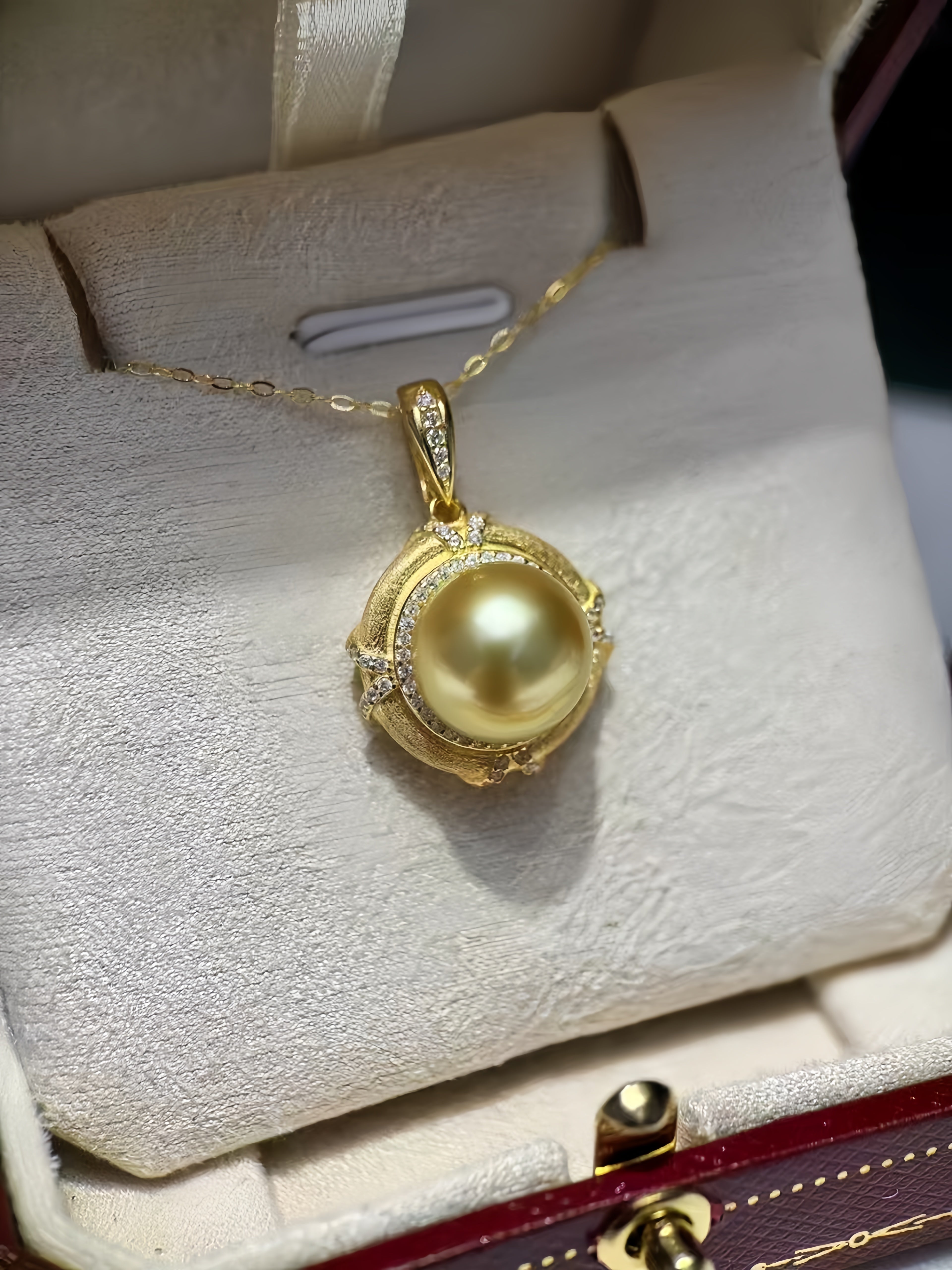 A gold pendant necklace featuring a large round yellow pearl surrounded by a decorative gold frame with small clear stones, displayed inside a cream-colored jewelry box.