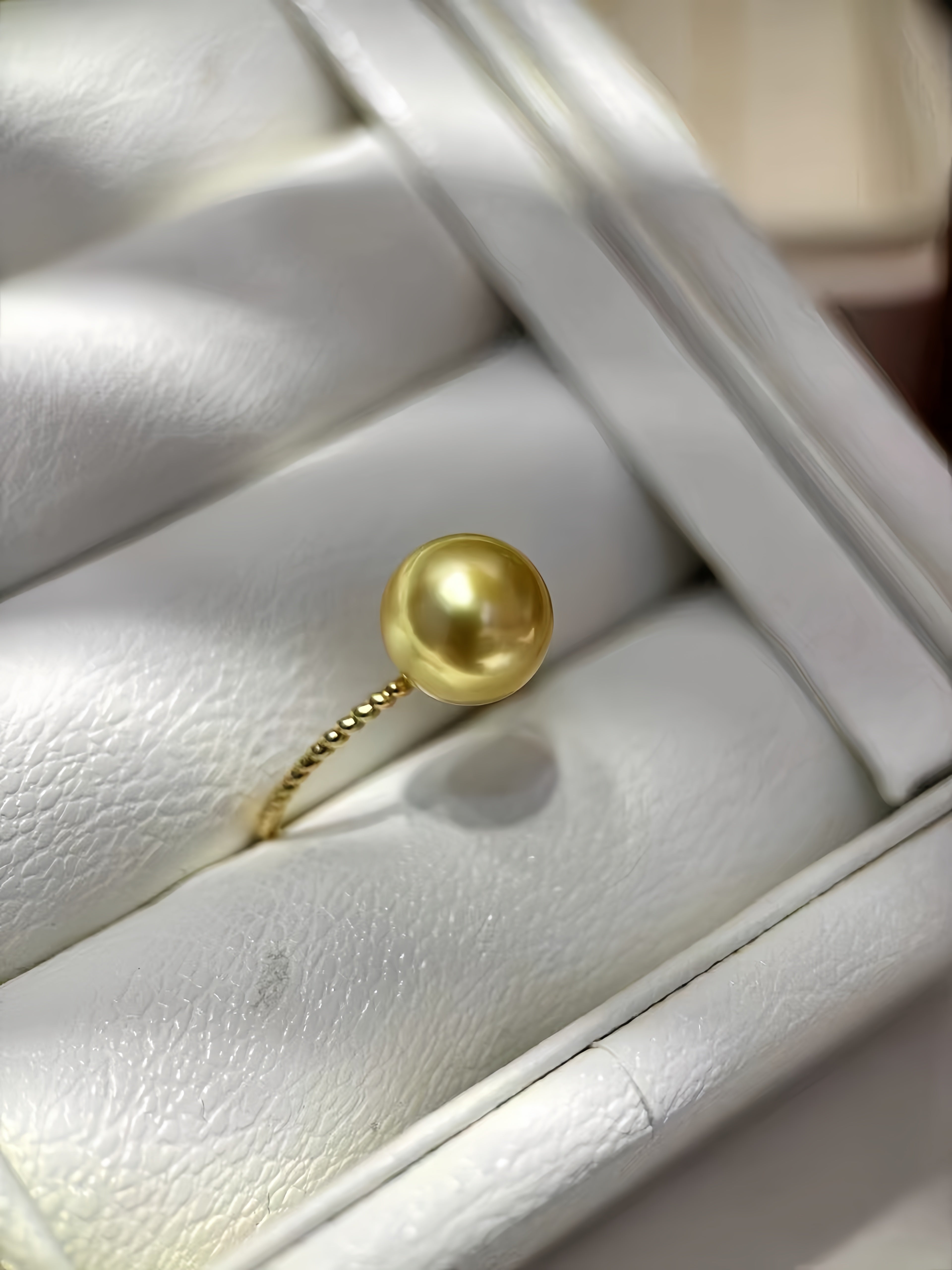 A gold ring featuring a round, lustrous pearl-like bead on a delicate beaded band, displayed inside a white textured jewelry box.