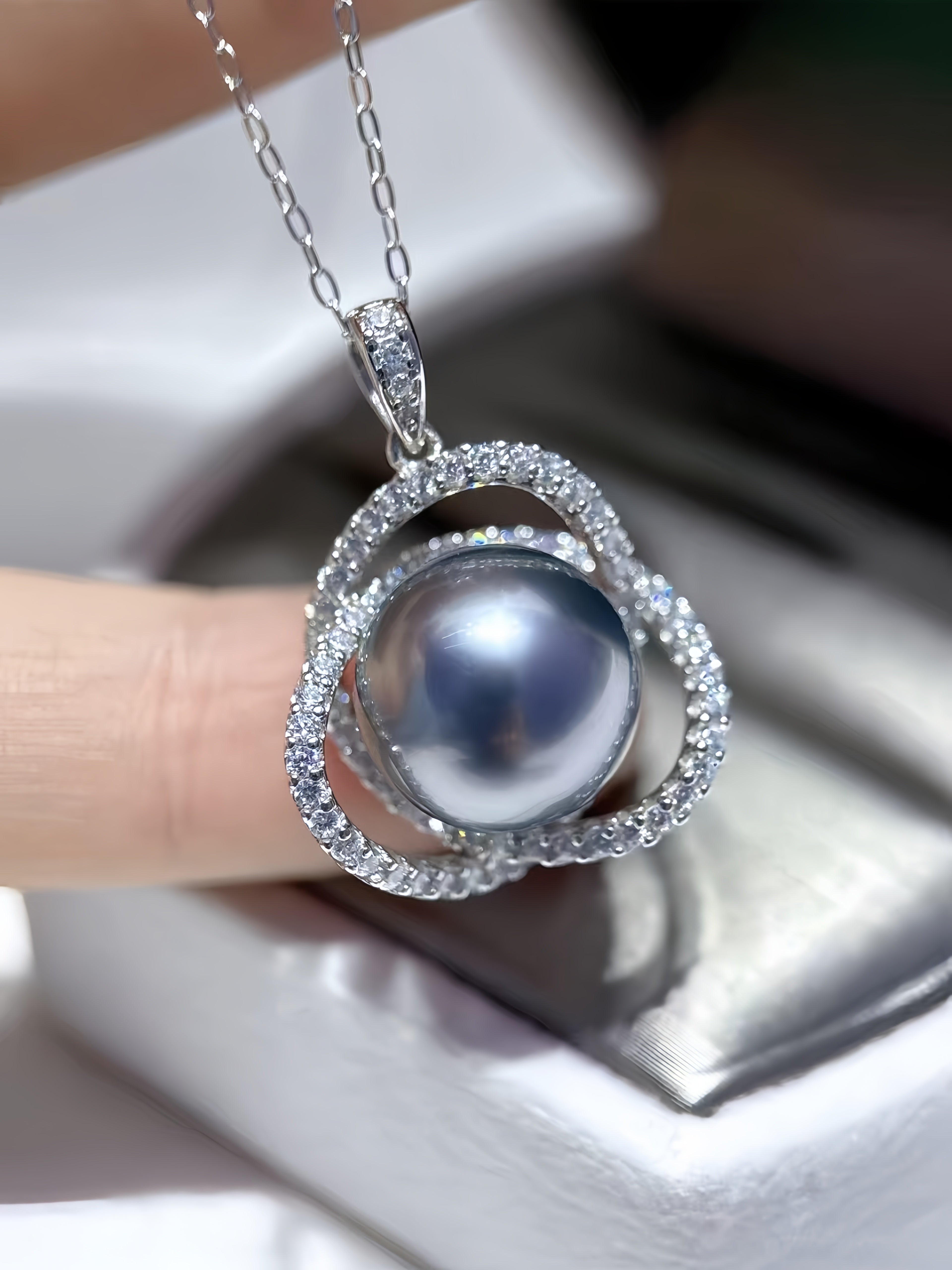 A close-up of a silver pendant necklace featuring a large round gray pearl surrounded by a double-ring halo of small clear stones, held by a hand over a white jewelry box.