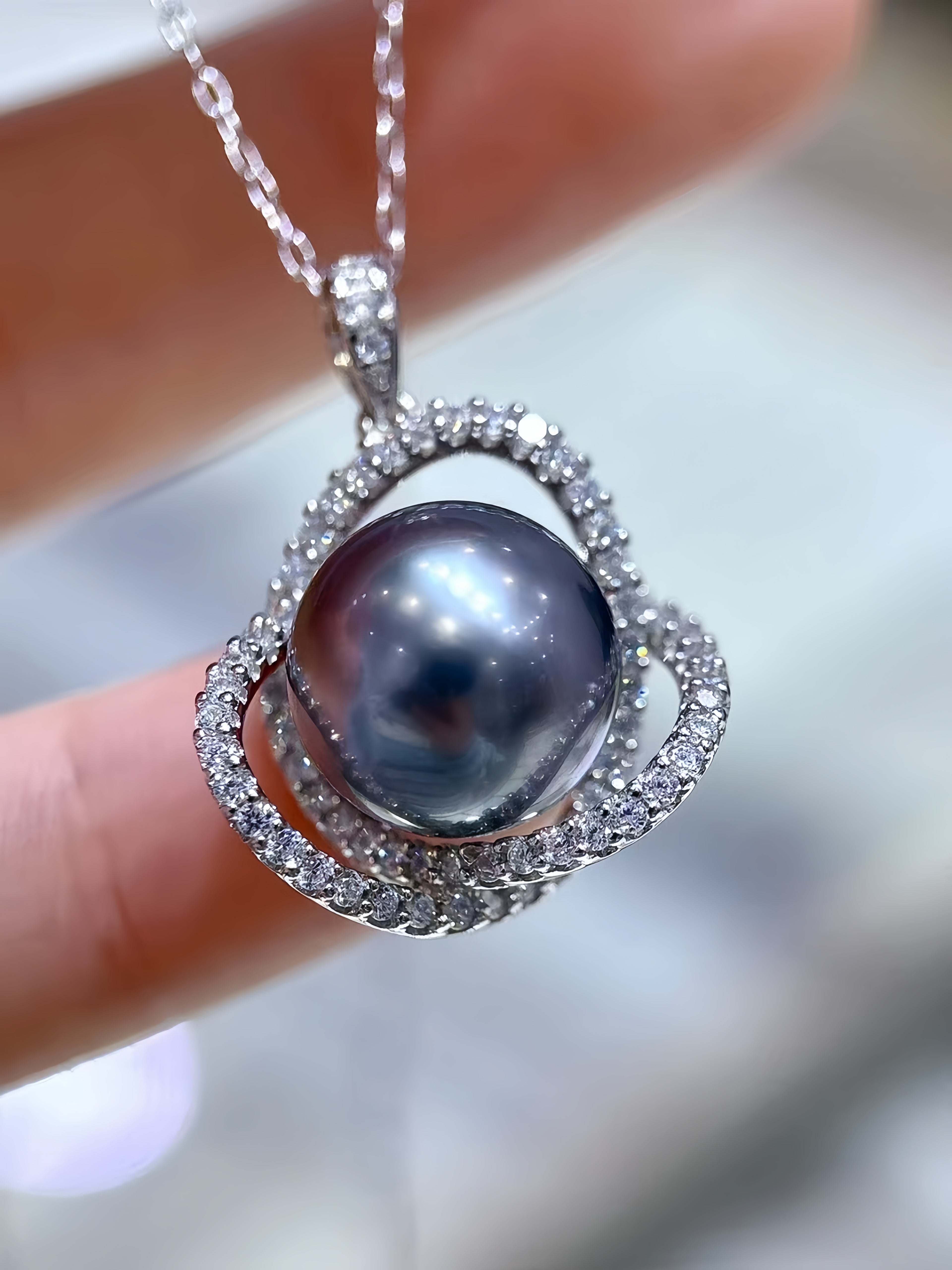 A close-up of a silver pendant necklace with a large oval dark pearl surrounded by small clear stones, held between fingers against a blurred light background.
