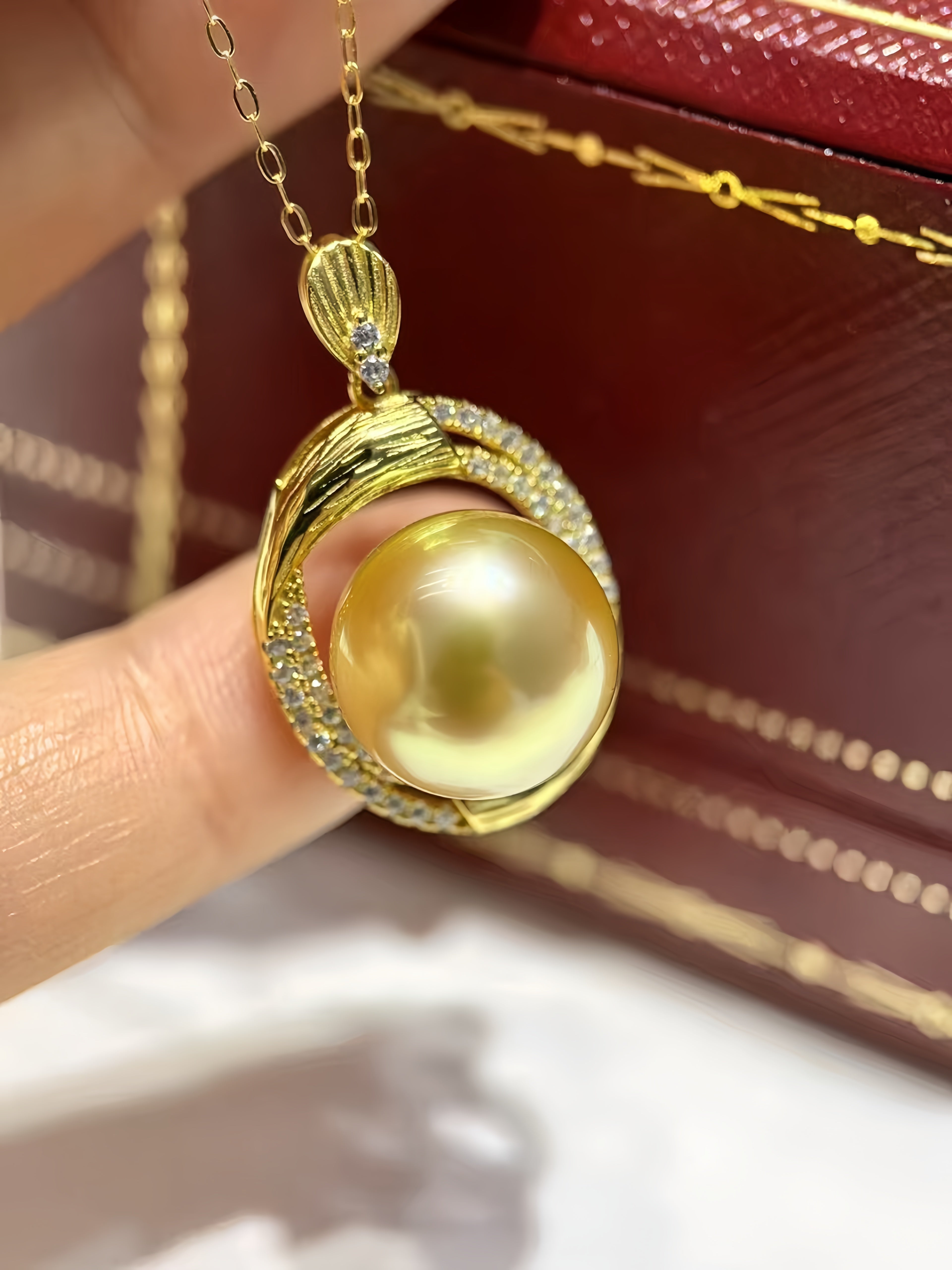 A gold pendant necklace with a large round yellow pearl and small clear stones, held between fingers with a dark red jewelry box in the background.