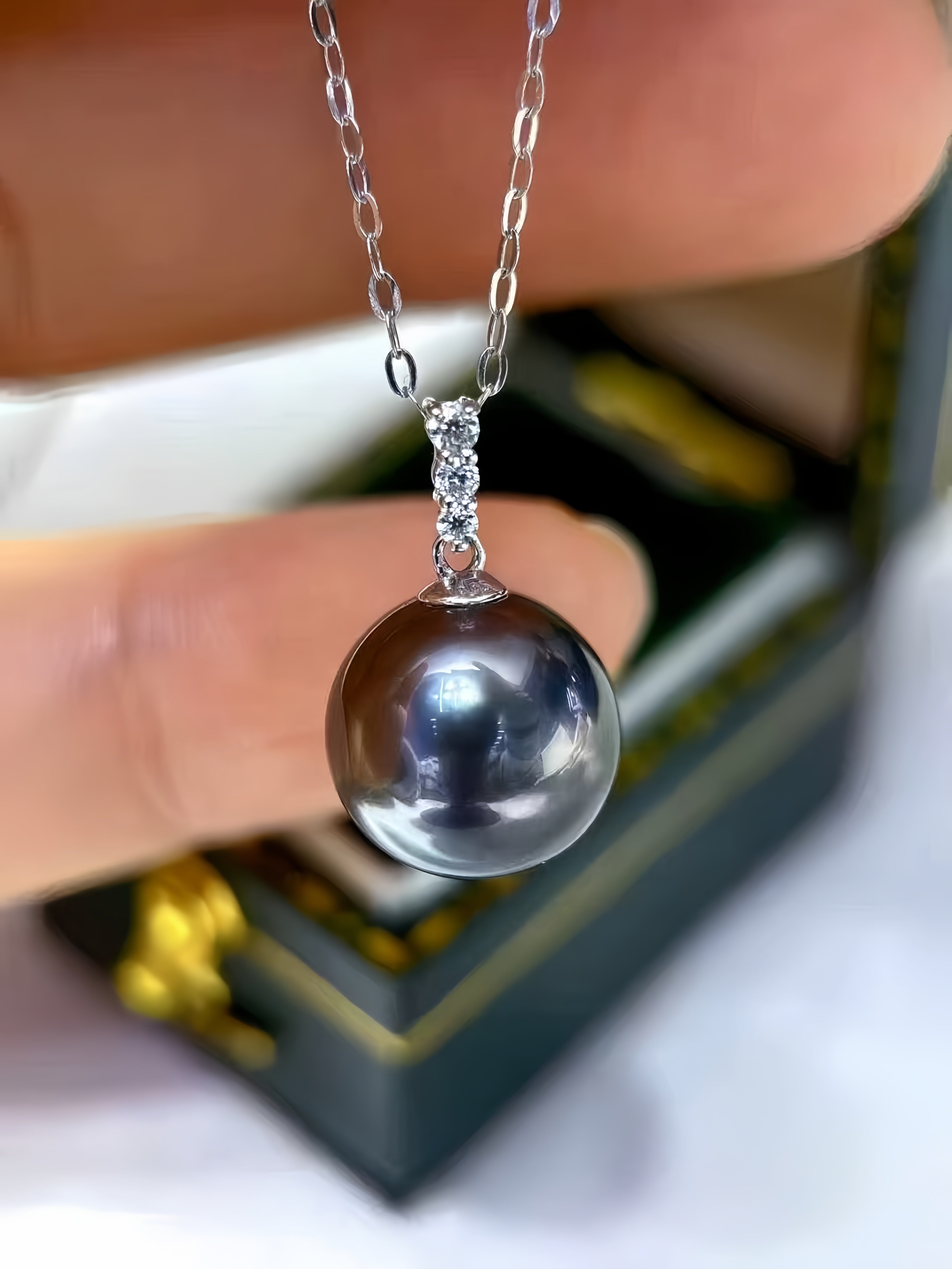 A close-up of a silver chain necklace with a round, dark gray pearl pendant held by fingers in front of a blurred jewelry box.