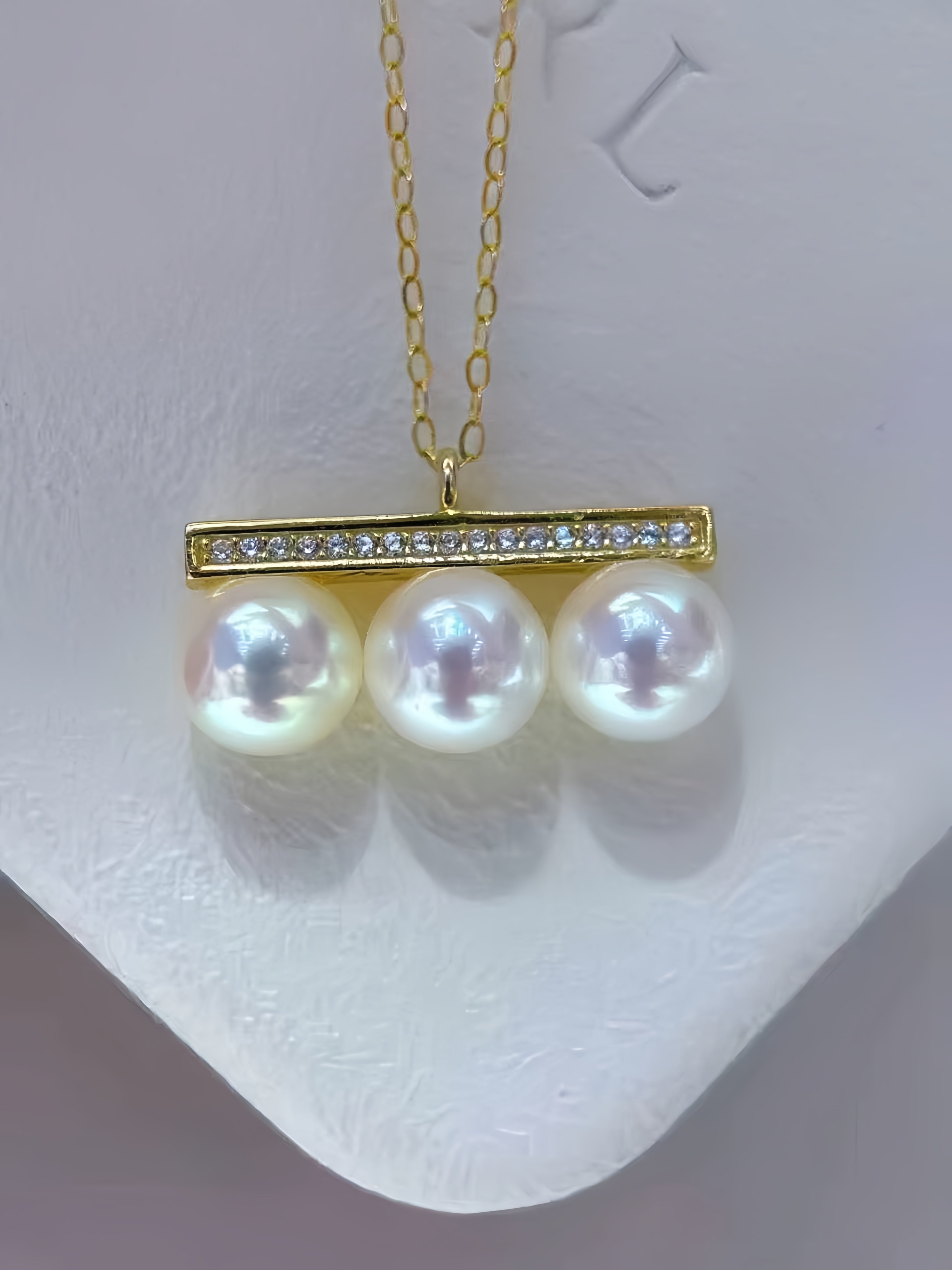 A gold pendant necklace featuring three large, lustrous white pearls hanging from a horizontal bar embellished with small clear stones, displayed on a white jewelry stand.
