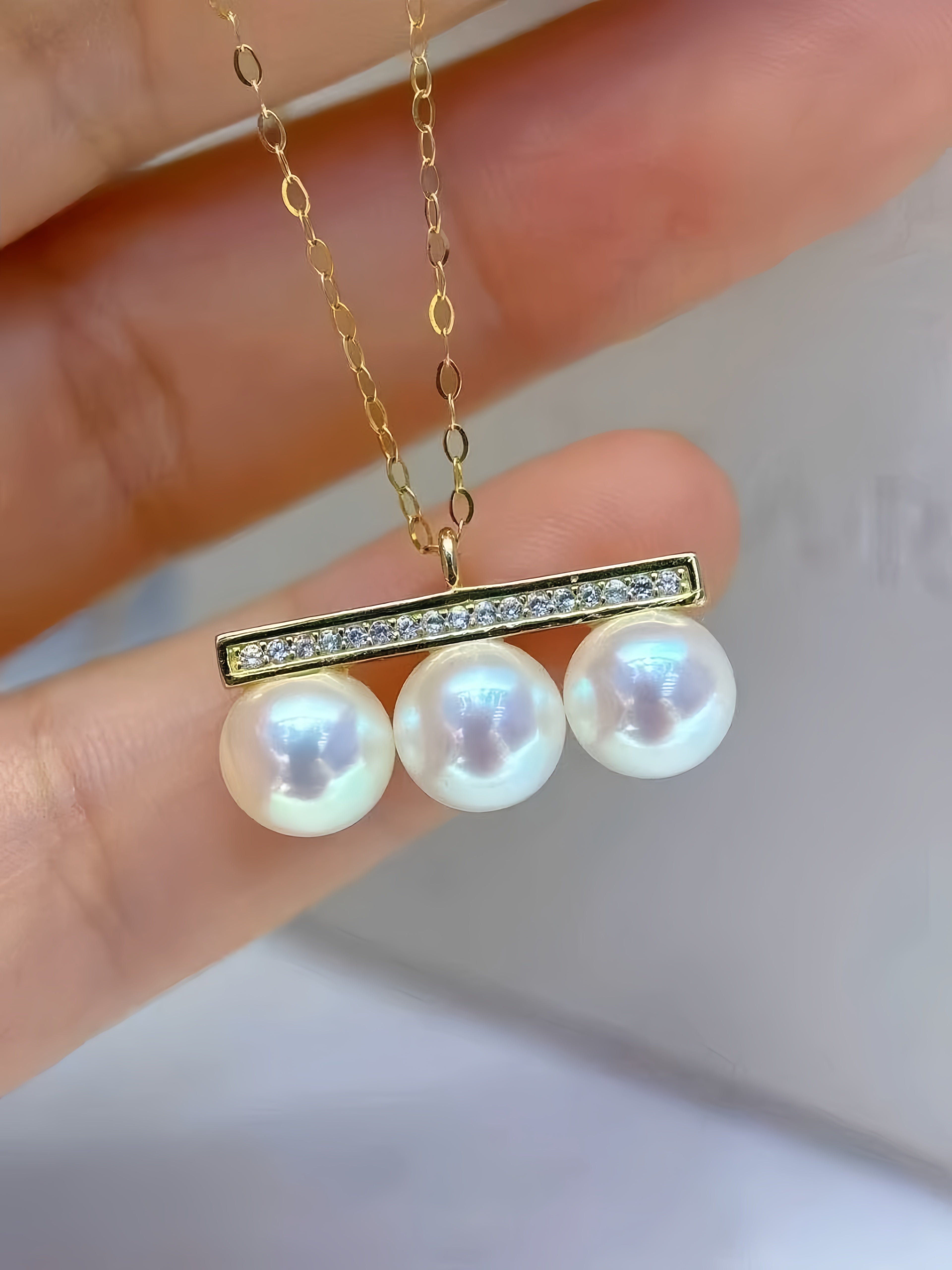 A gold necklace with a bar pendant featuring three white freshwater pearls and a row of small clear stones, held between fingers.