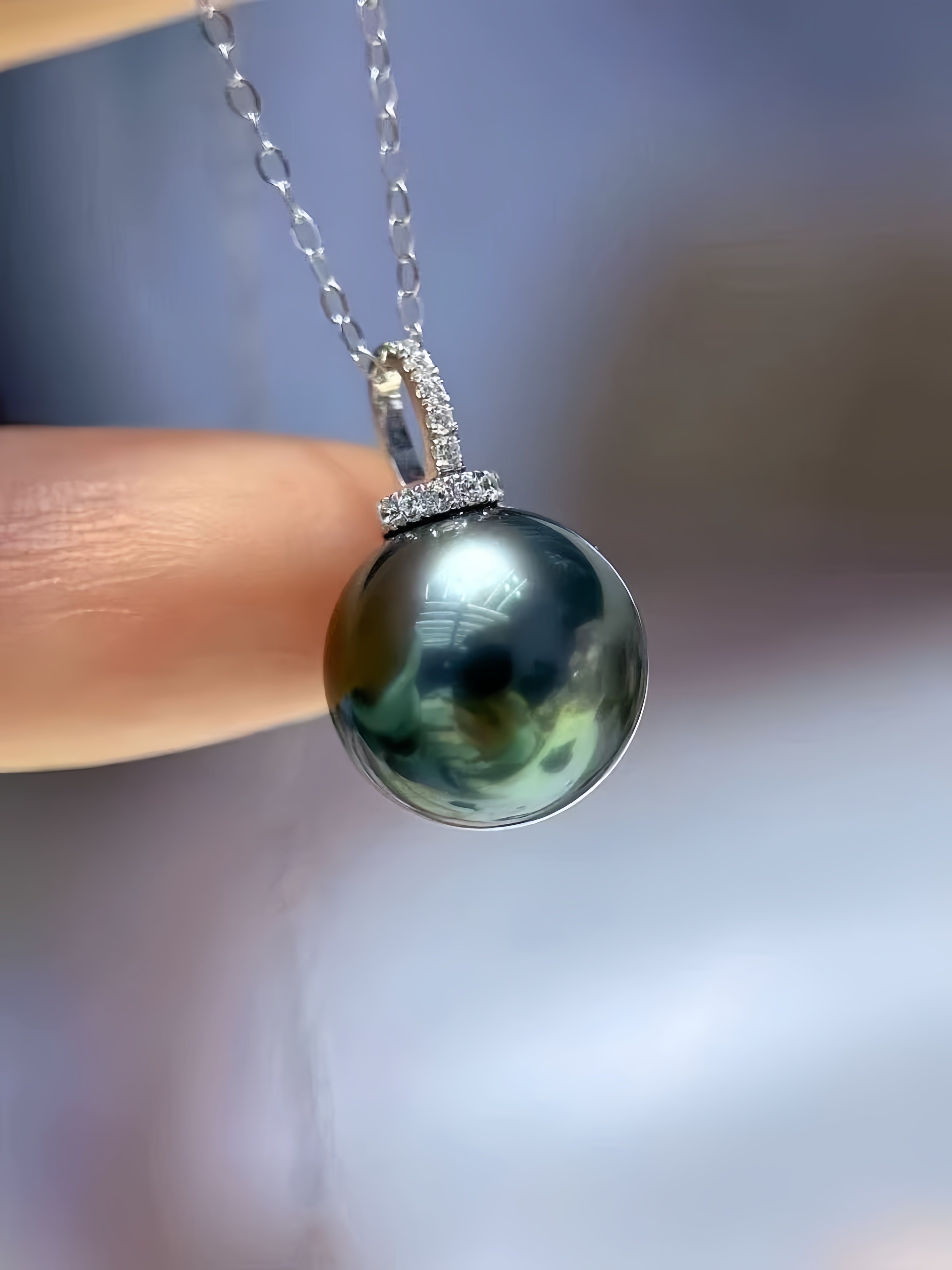 A close-up of a dark green-black pearl pendant necklace with a silver-toned setting, held by a person's finger against a soft blurred background.