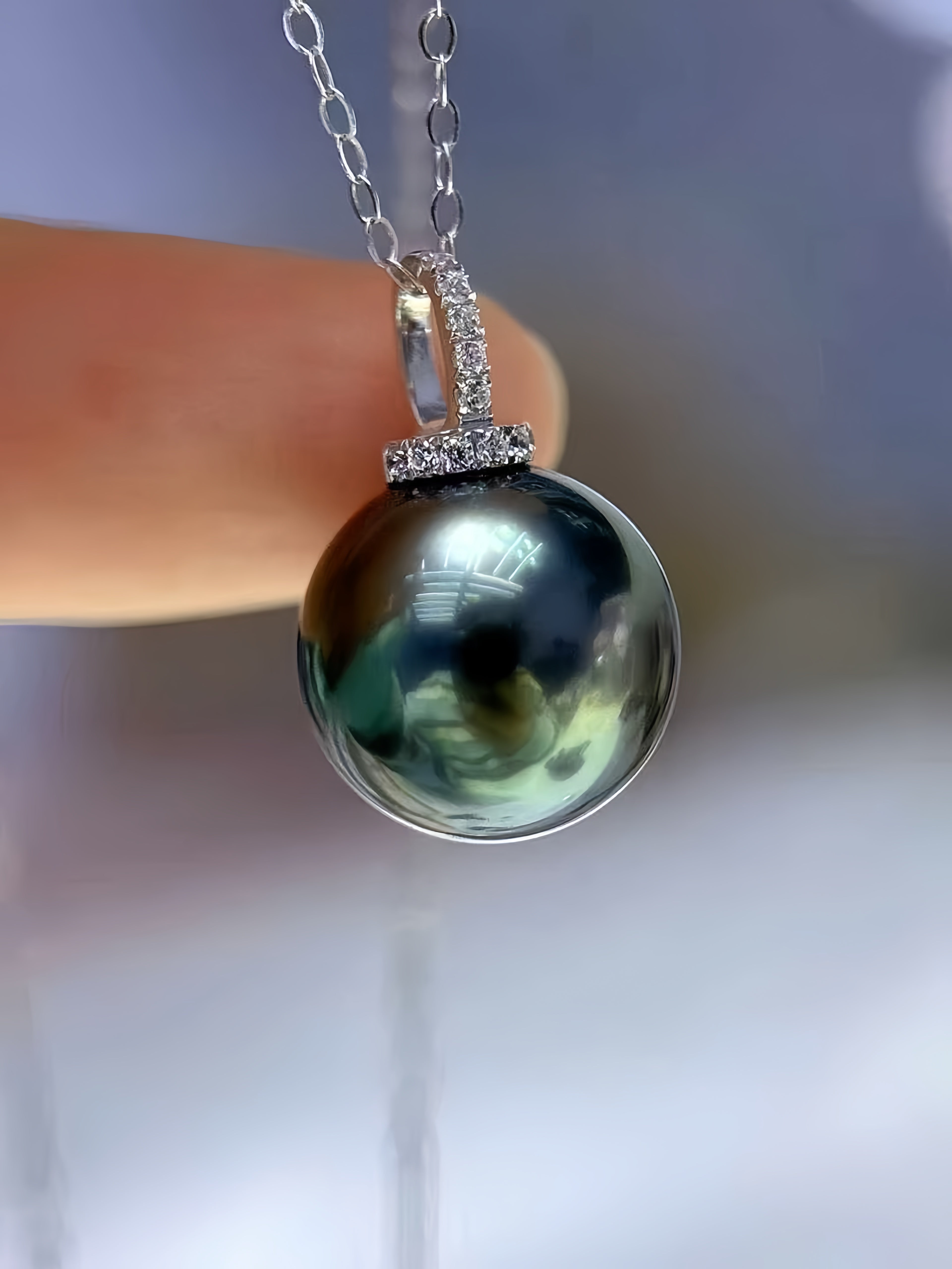 A close-up of a silver pendant necklace with a large, round, dark green-black pearl and small clear stones, held by a finger against a soft, blurred background.