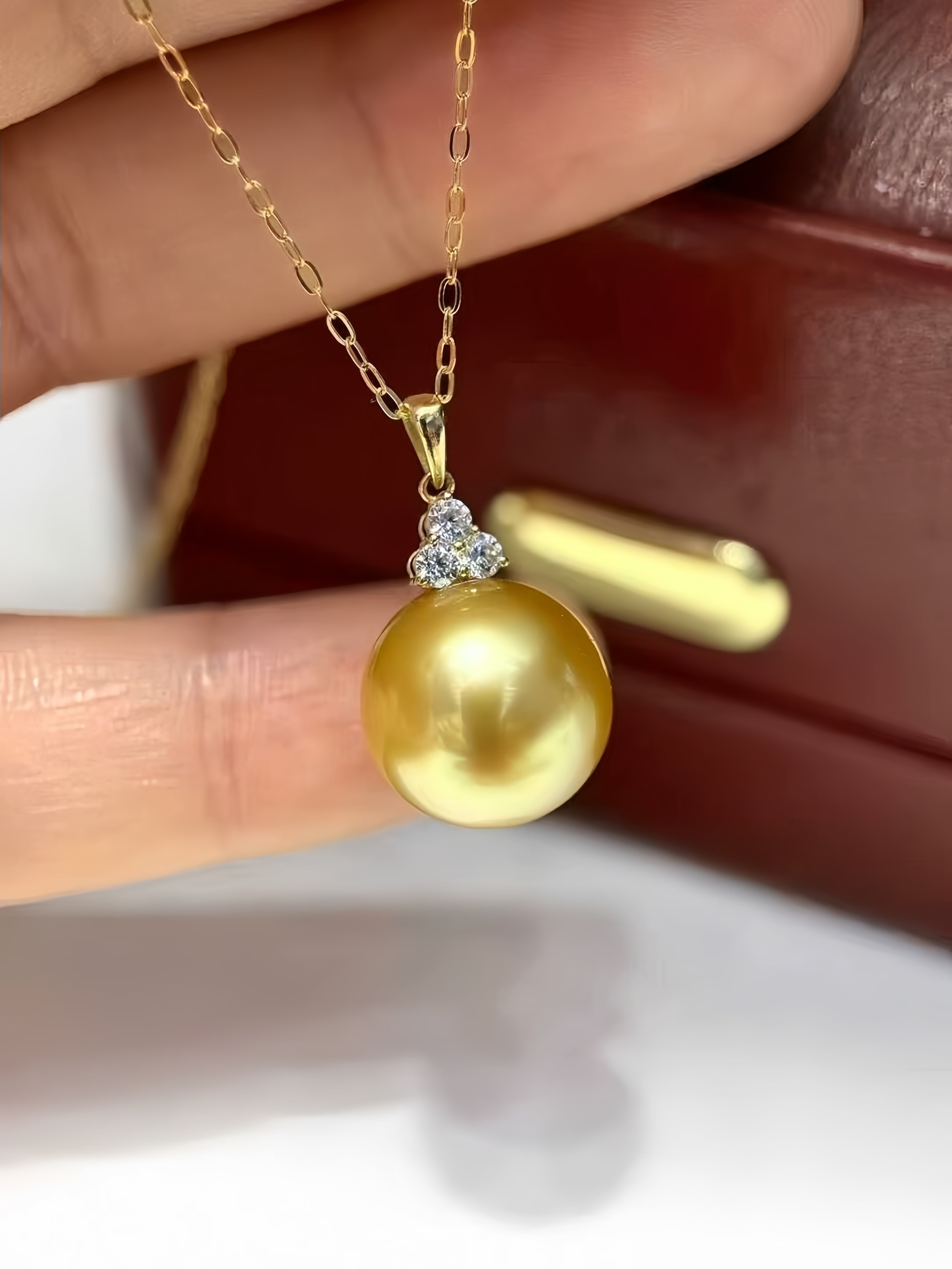 A gold pendant necklace featuring a large, lustrous yellow pearl accented by a small cluster of clear stones, held by a hand against a dark brown leather background.