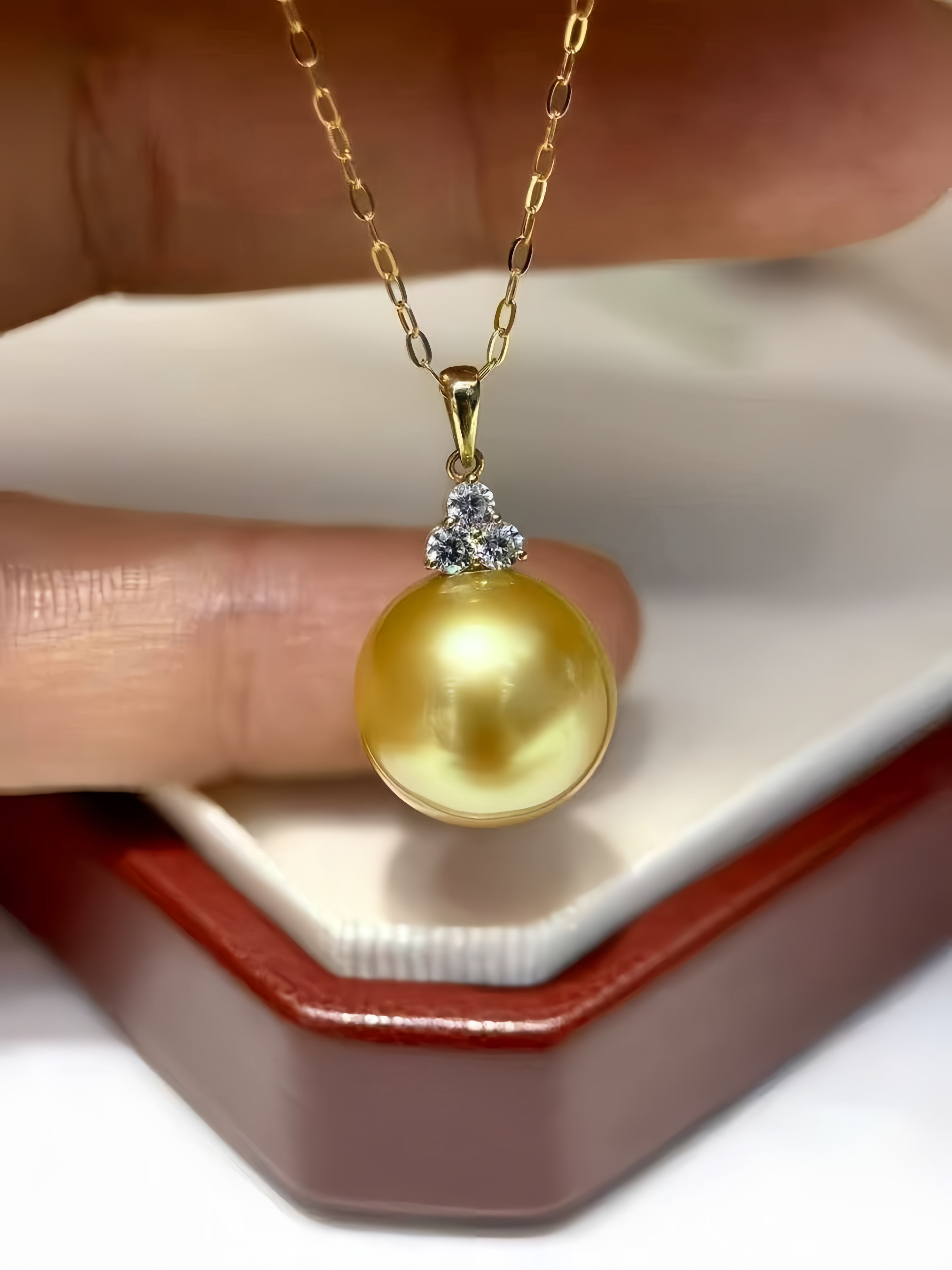 A gold pendant necklace featuring a large round yellow pearl accented by a small cluster of clear stones, held by a hand above an open red and white jewelry box.