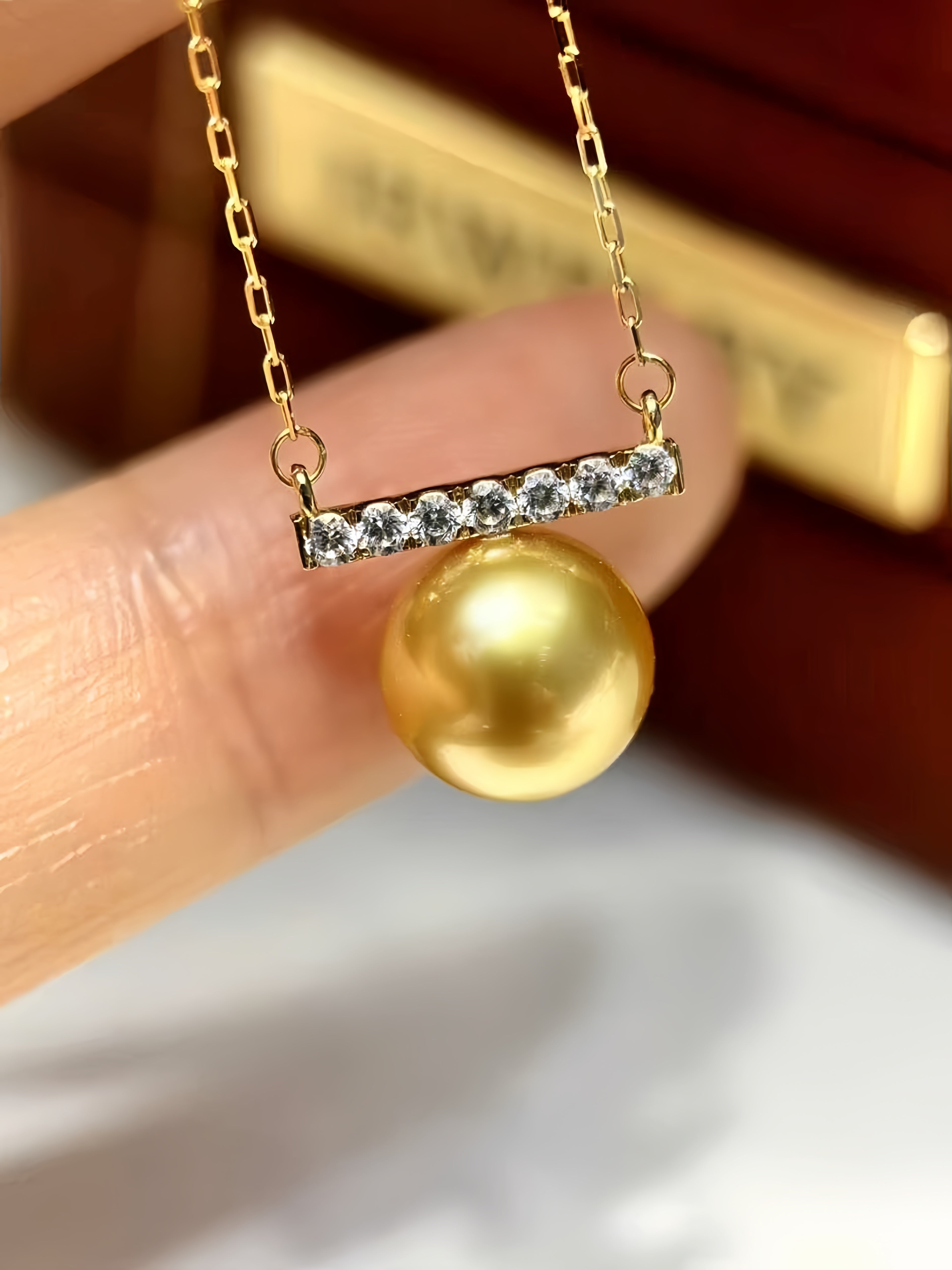 A gold pendant necklace featuring a bar set with small clear stones and a large round gold pearl, held between fingers against a blurred dark brown jewelry box background.