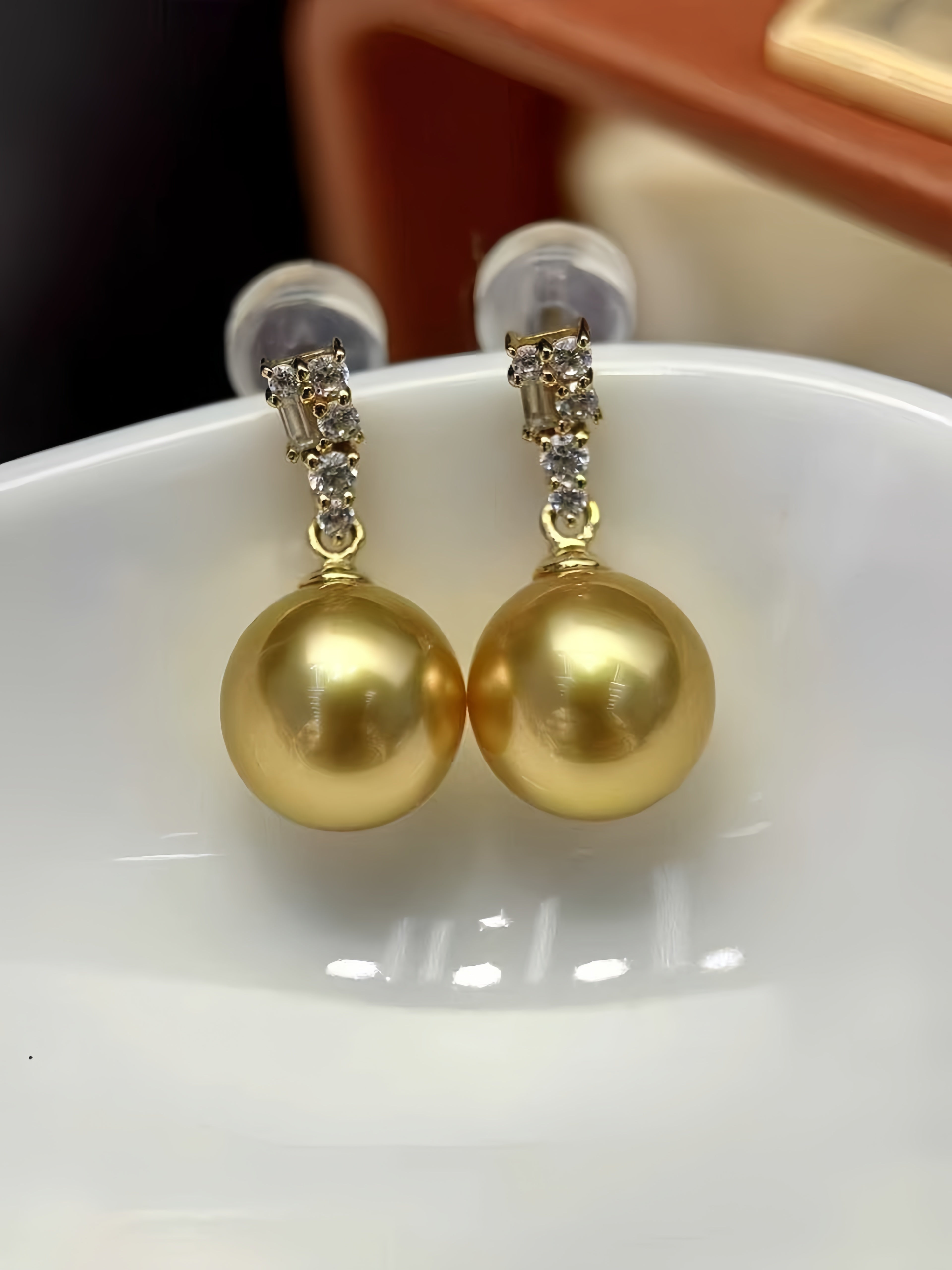 A pair of gold drop earrings with large round gold pearls and small clear crystal accents at the top, displayed on a white ceramic dish.