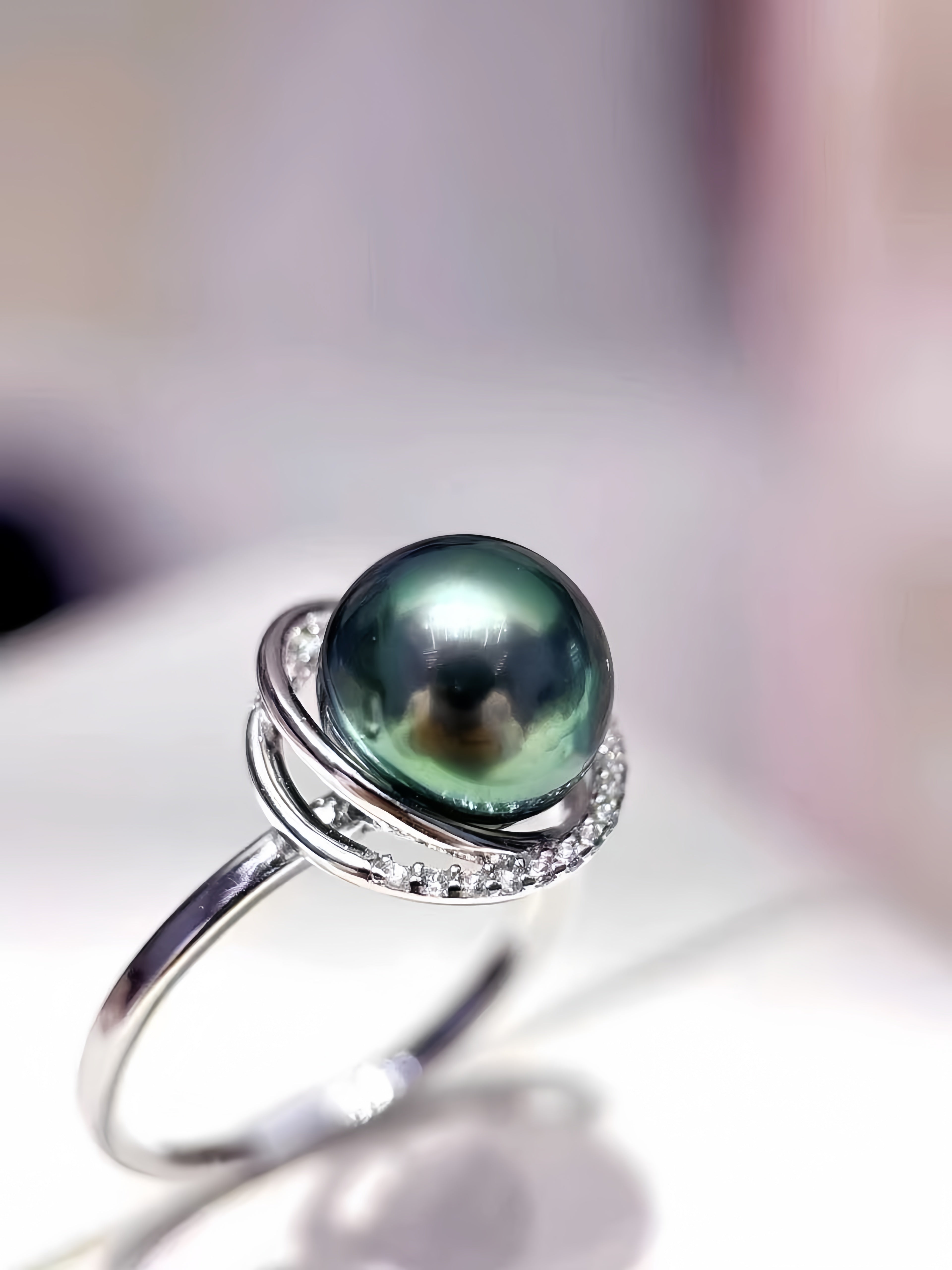 A silver ring featuring a large, round, dark green-black pearl with iridescent highlights, accented by a halo of small clear stones on a soft, blurred light background.