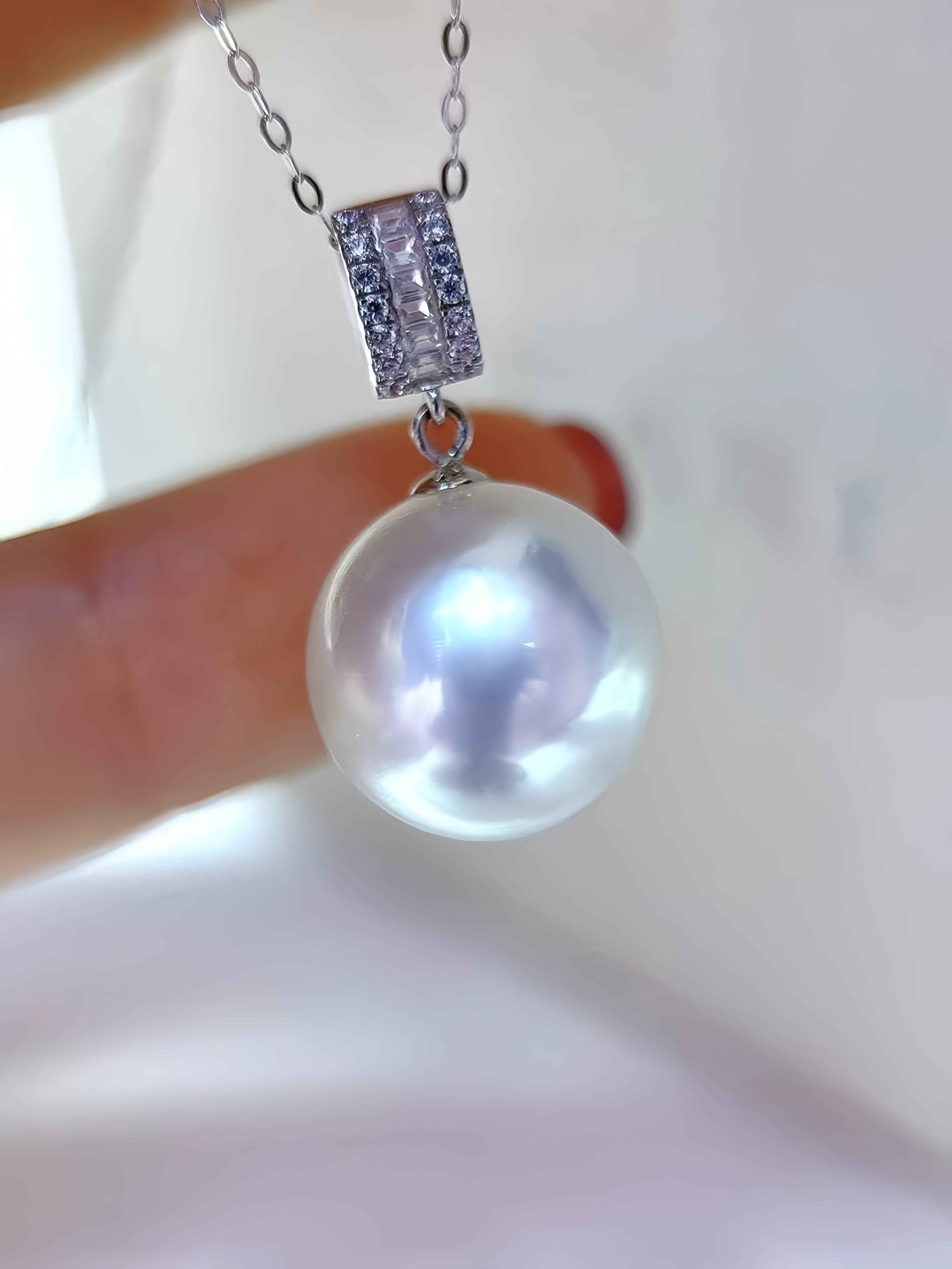 A close-up of a silver pendant necklace featuring a large, lustrous white pearl and a rectangular pavé-set bezel, held by a finger against a soft, out-of-focus white background.