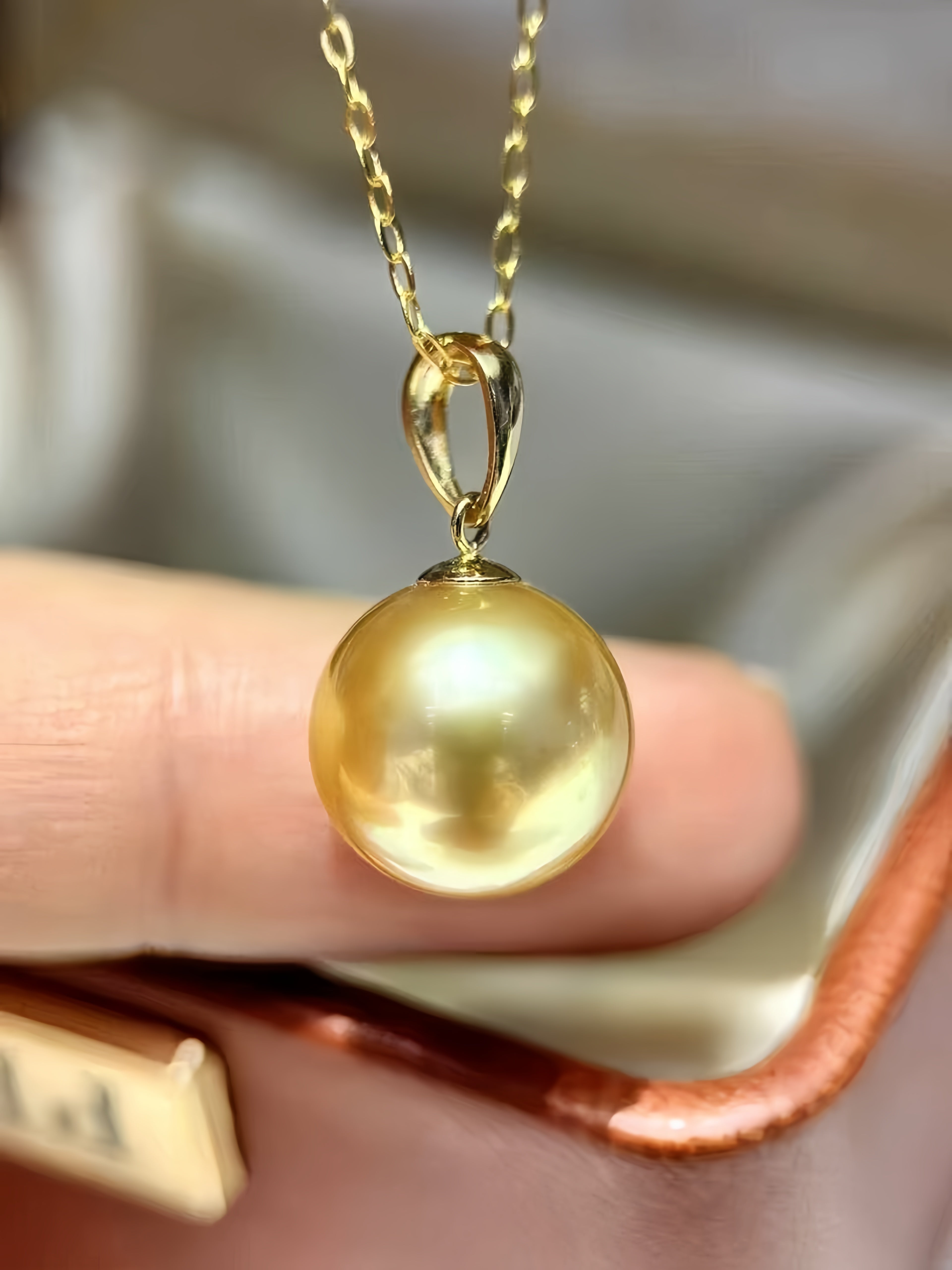 A close-up of a gold pendant necklace featuring a large, lustrous round pearl pendant, held by a person's fingers over a jewelry box.