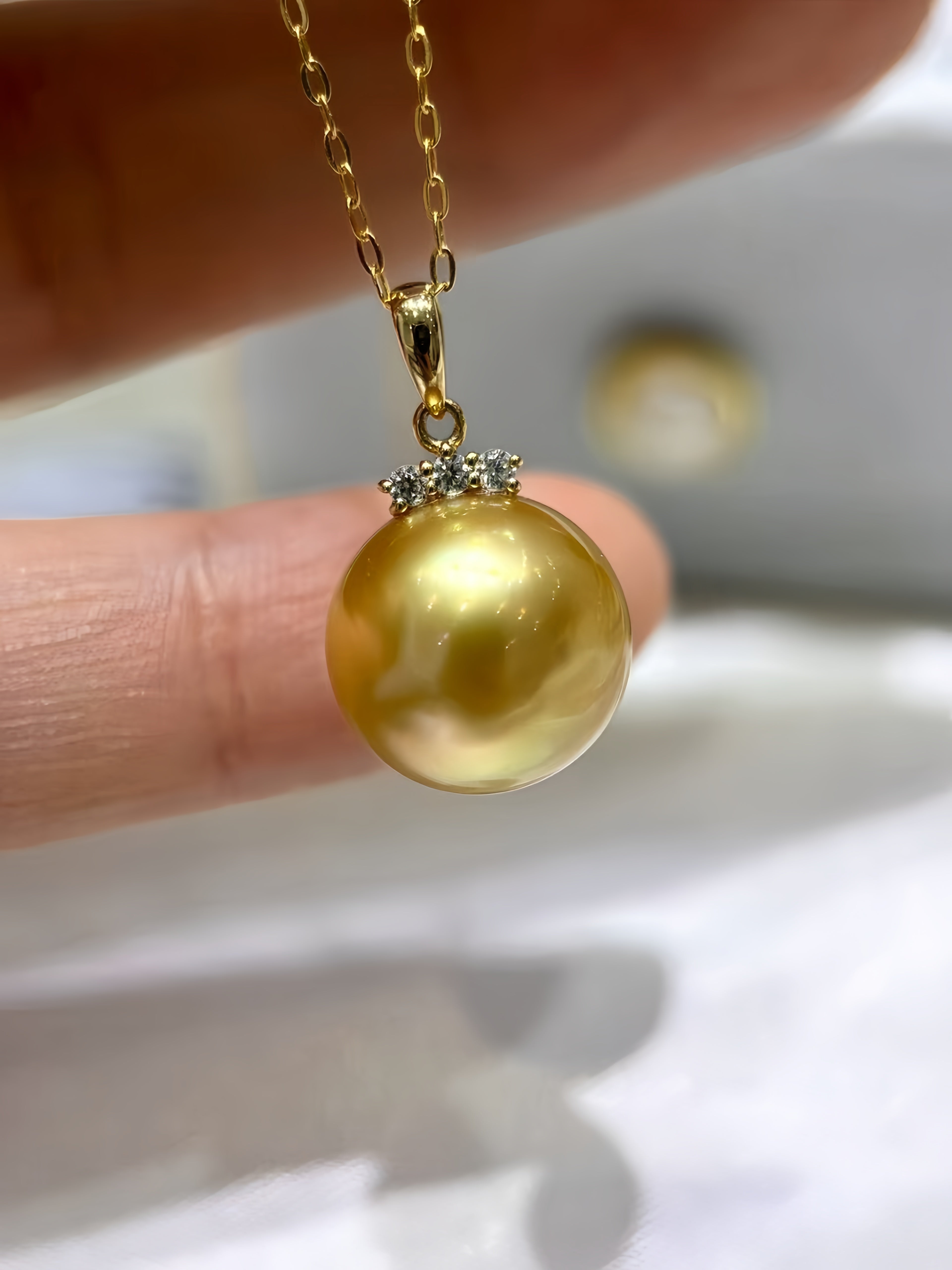 A close-up of a gold pendant necklace featuring a large, lustrous gold pearl pendant with a small cluster of clear stones at the top, held by fingers.