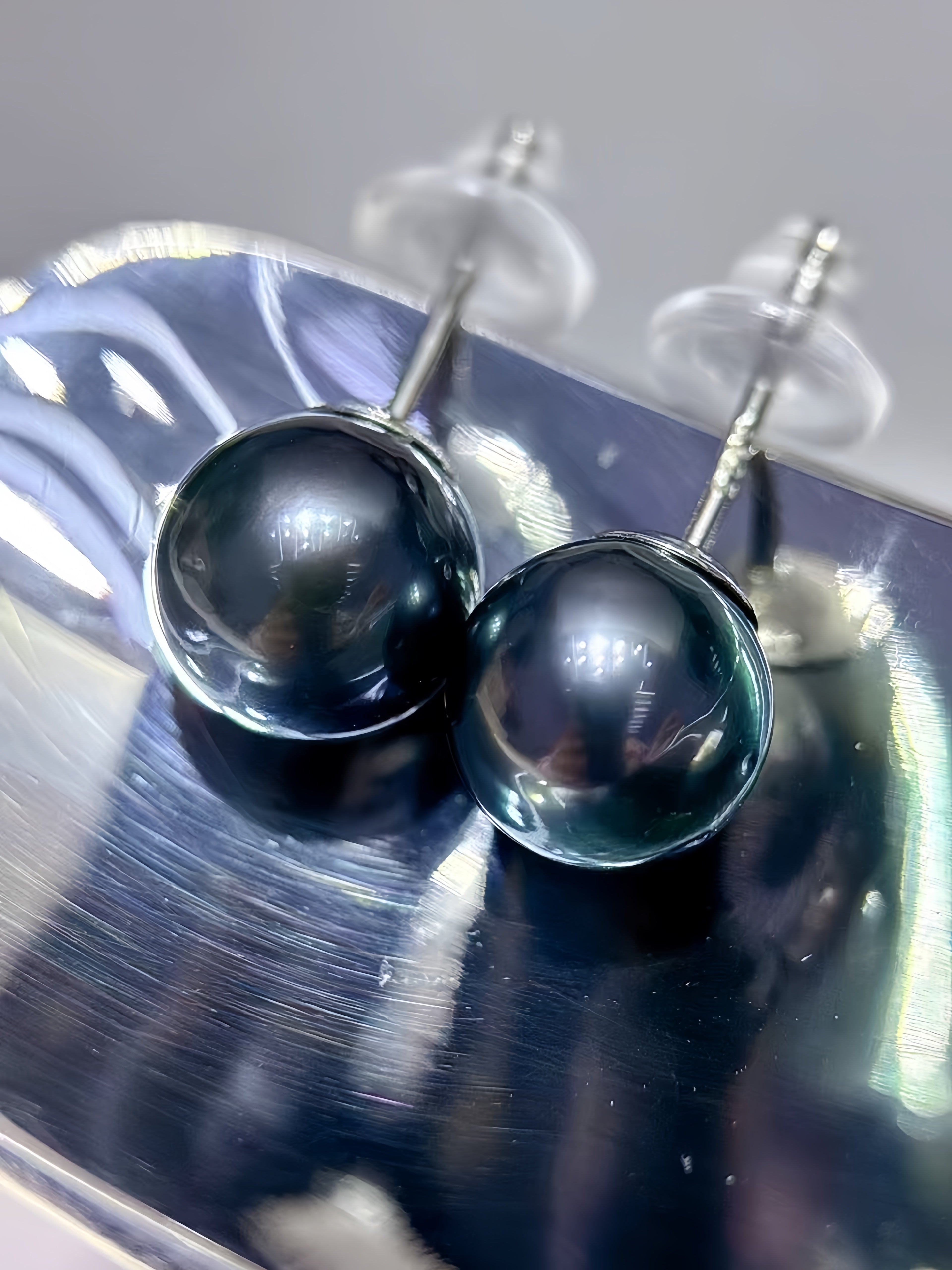 A pair of round black pearl stud earrings with silver posts and clear butterfly backings, resting on a reflective silver tray.