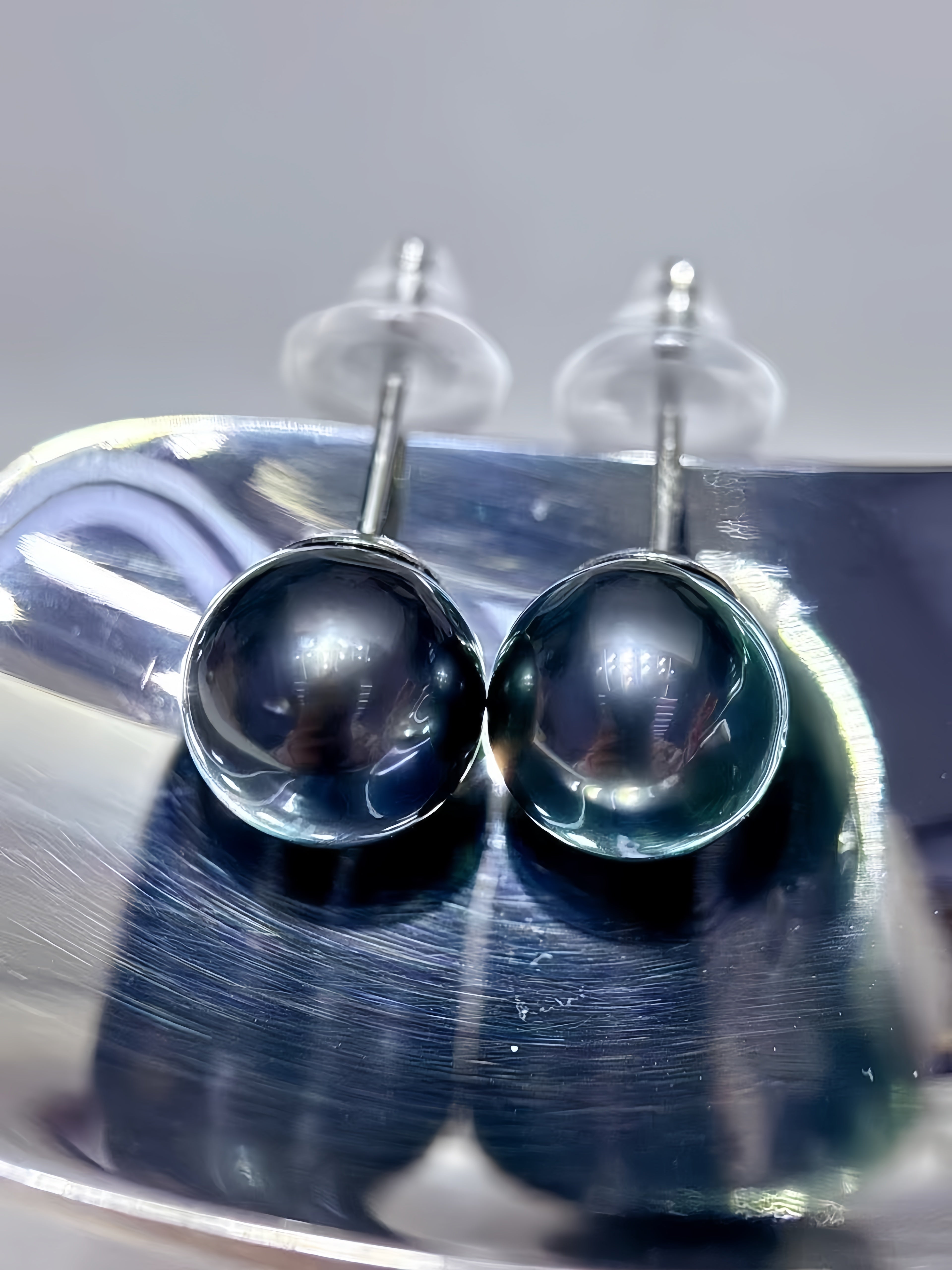 A pair of round black pearl stud earrings with silver posts and clear butterfly backings, resting on a reflective silver metal surface.