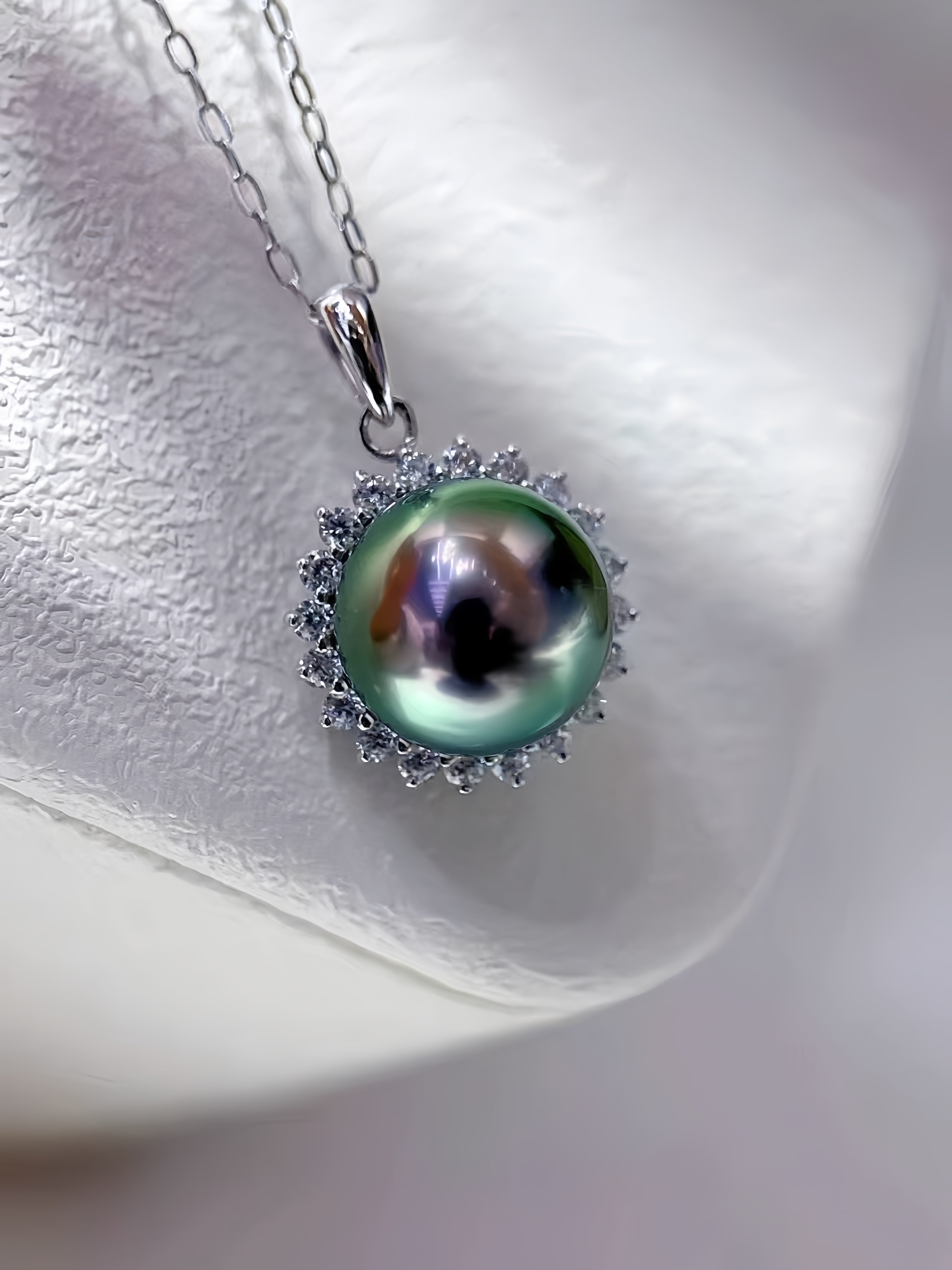 A silver pendant necklace featuring a large, iridescent green pearl surrounded by a halo of small clear stones, resting on a textured white display stand.
