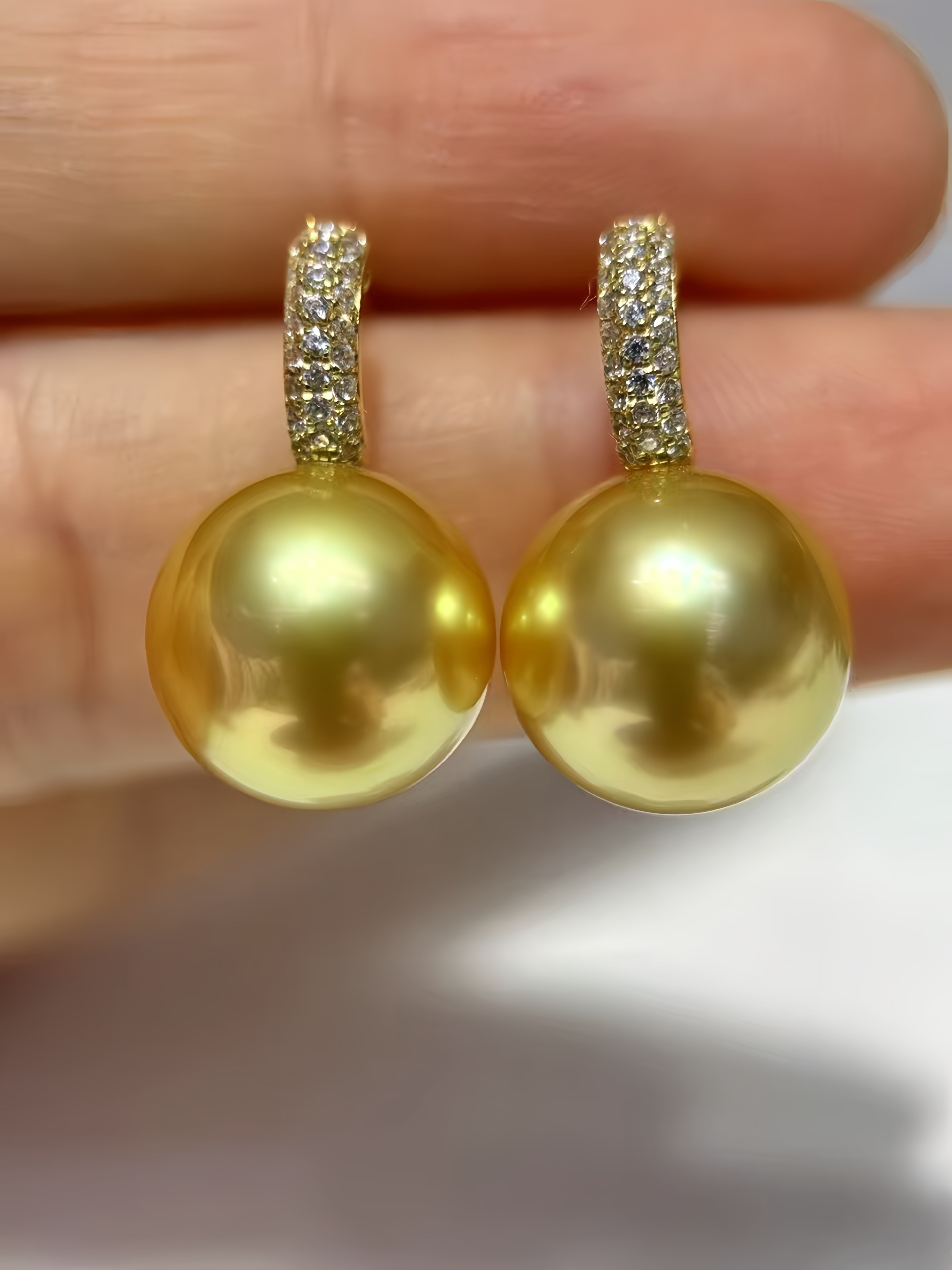 A pair of gold-toned drop earrings with round yellow-gold pearls and pavé-set clear stones at the top, shown resting on a person's fingers against a soft light background.