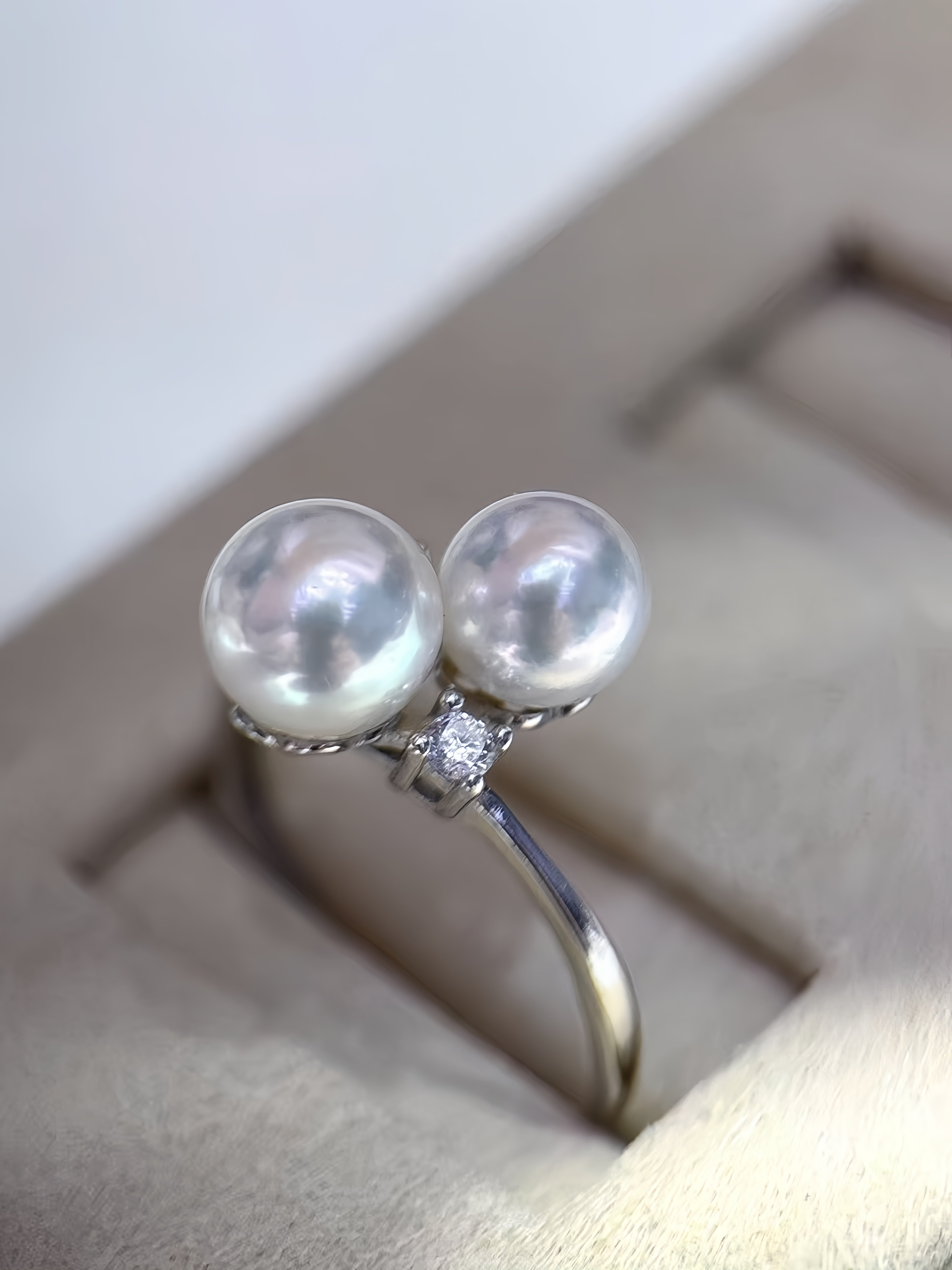 A silver ring featuring two large, lustrous white pearls and a small clear diamond accent, displayed in a beige jewelry box.