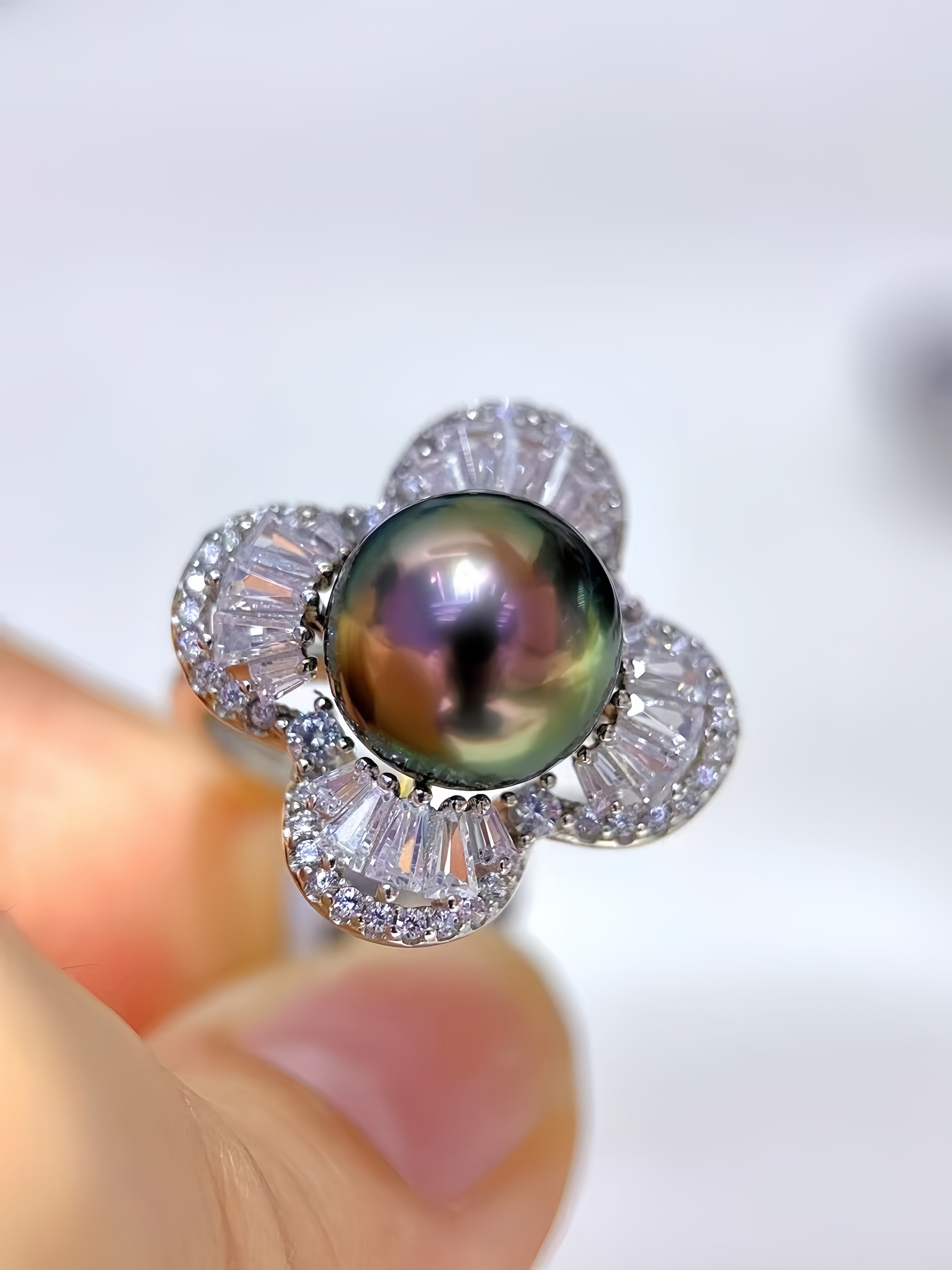 A close-up of a flower-shaped ring featuring a large iridescent dark pearl in the center, surrounded by five clear baguette and round stones in a silver-toned setting, held by fingers against a soft white background.