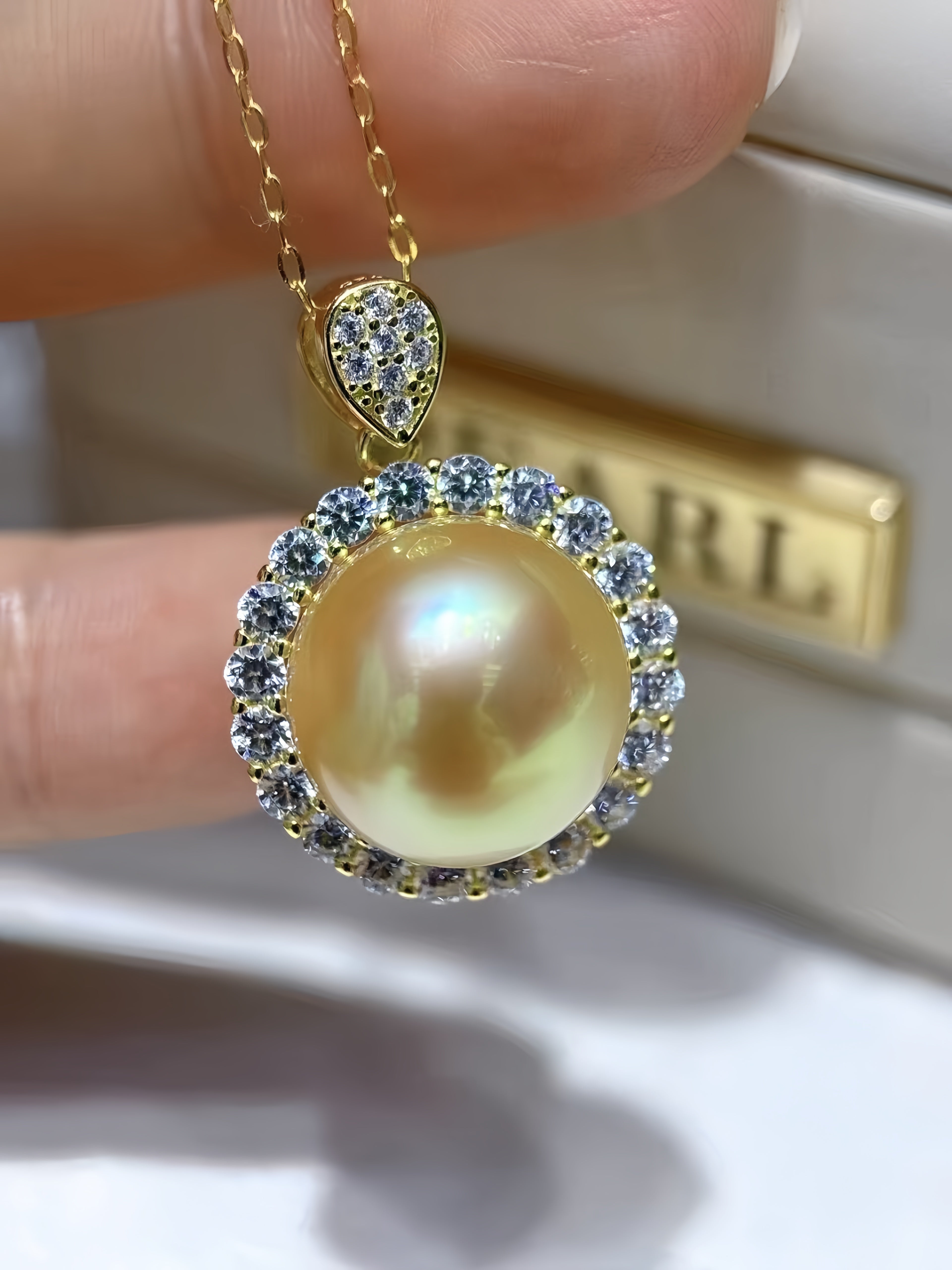 A gold pendant necklace with a large iridescent pearl surrounded by small clear stones, held between fingers with a blurred jewelry box in the background.