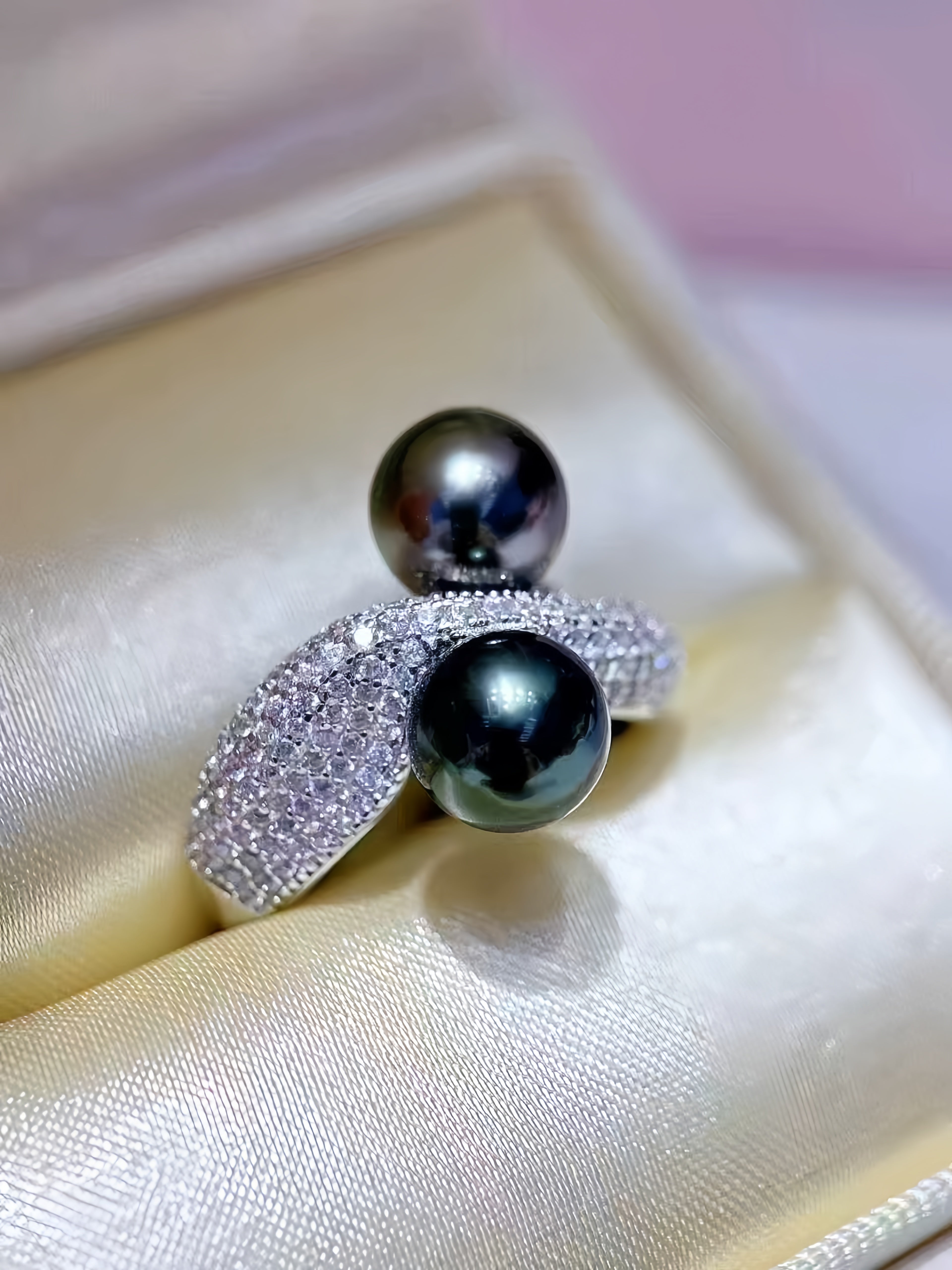 A close-up of a silver ring with two large dark pearls and pavé-set clear stones, displayed on a cream satin ring box.