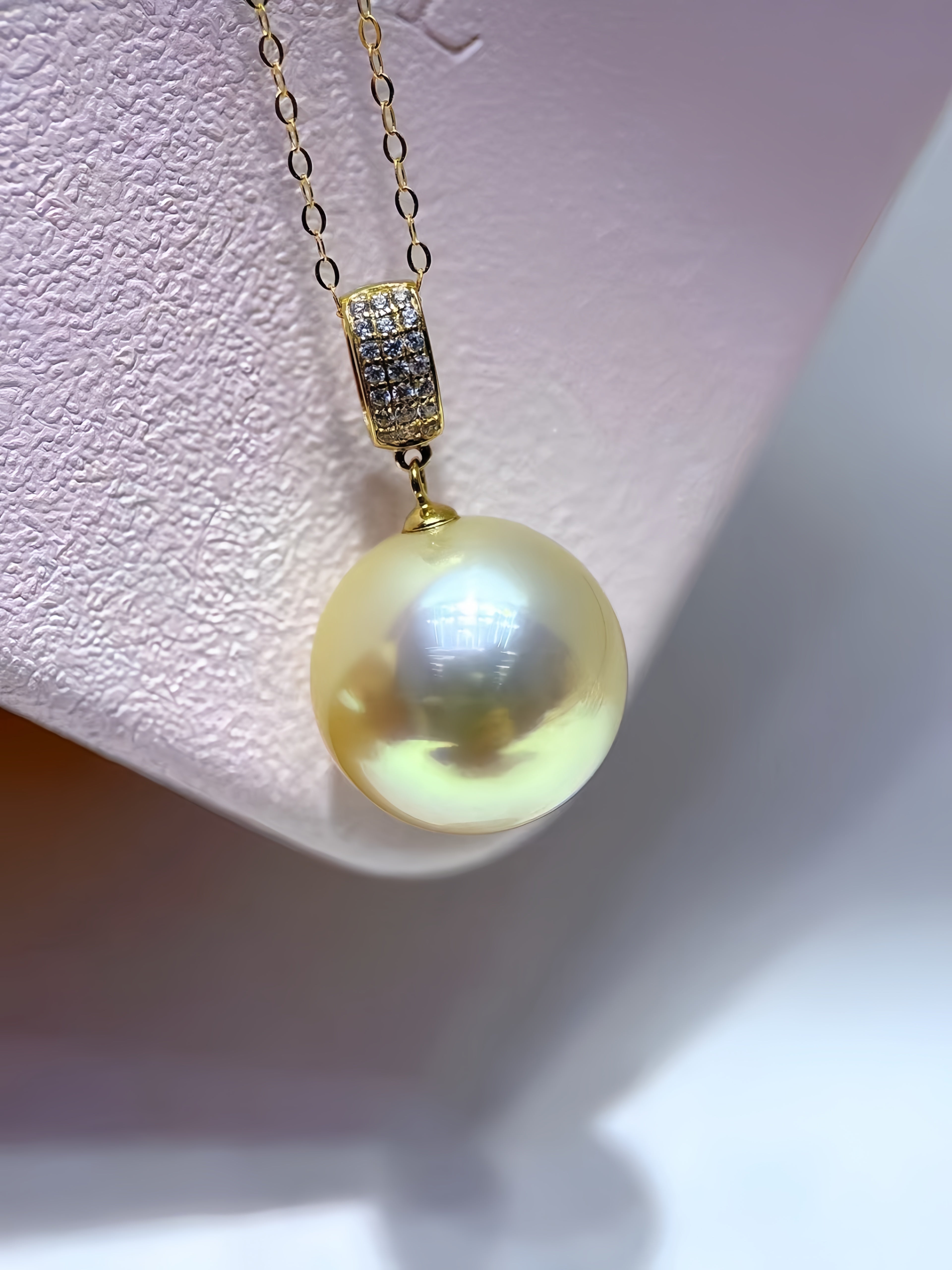 A gold pendant necklace featuring a large, lustrous yellow pearl and a small pavé-set gold accent, displayed on a textured light pink jewelry stand.