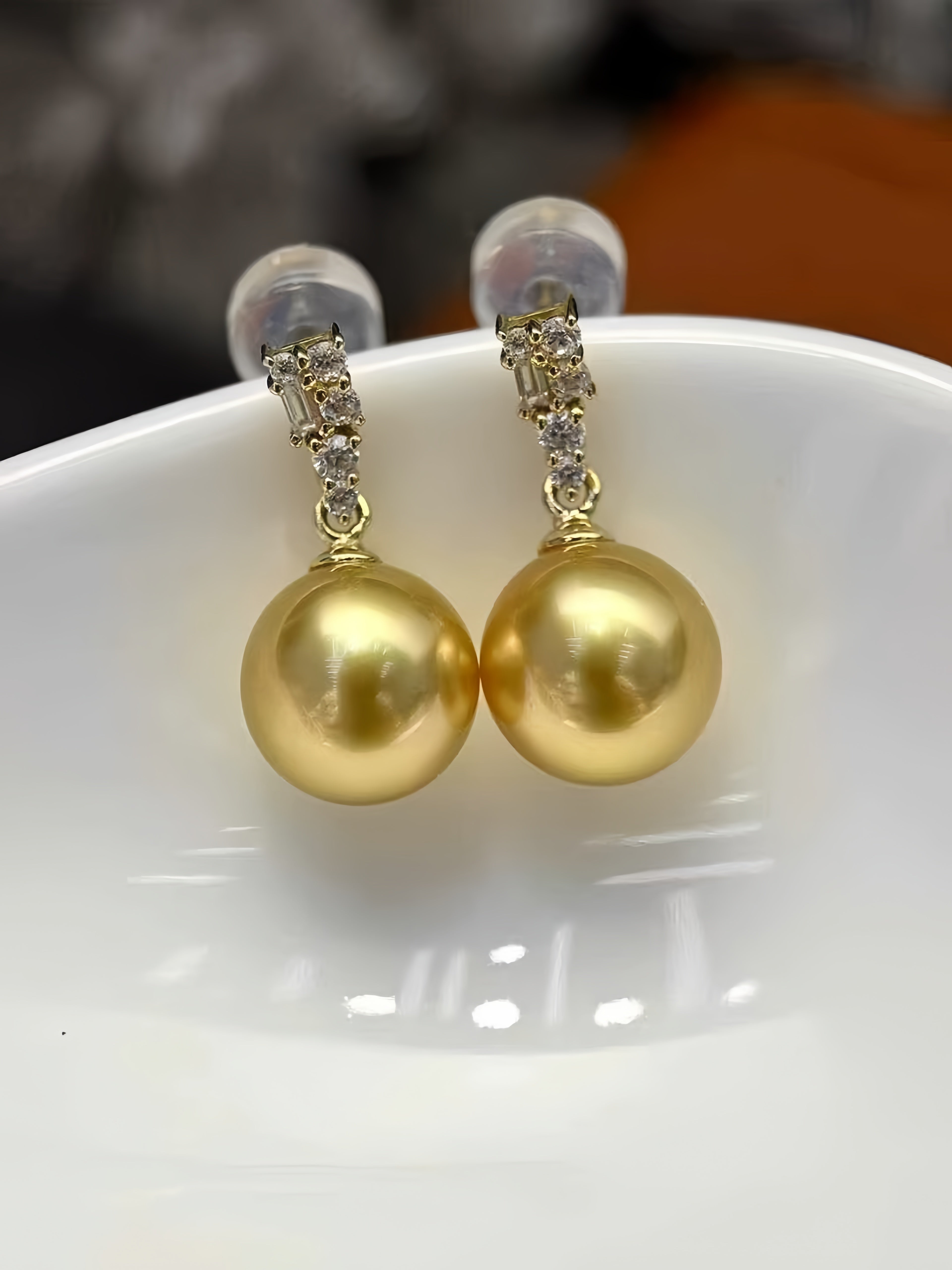 A pair of gold drop earrings with large round gold pearls and small clear stone accents at the top, displayed on a white ceramic dish.