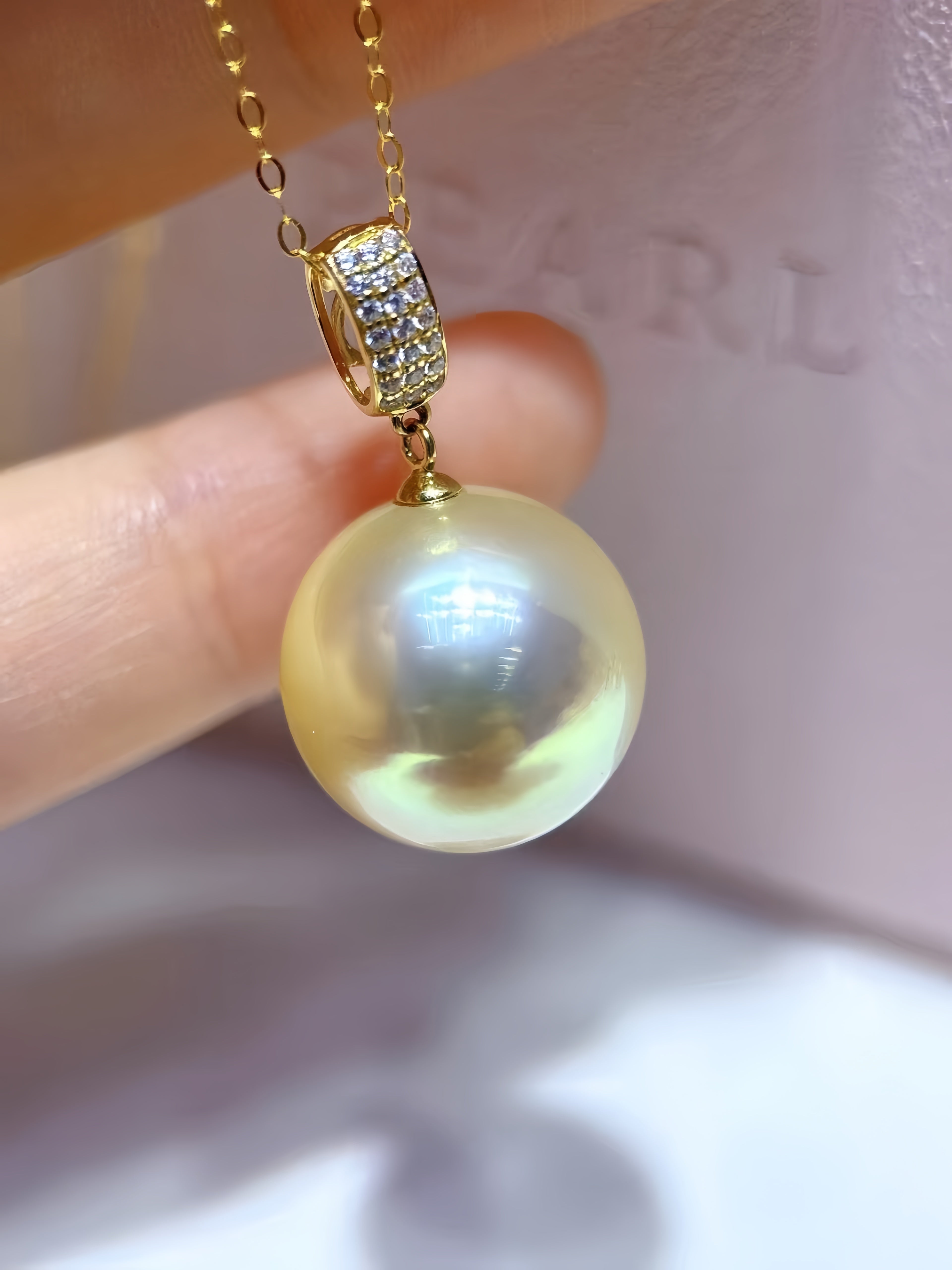 A close-up of a hand holding a gold necklace with a large round pearl pendant and a gold bezel-set crystal accent. The background is a soft-focus light pink surface with the brand name 'CARL' faintly visible.