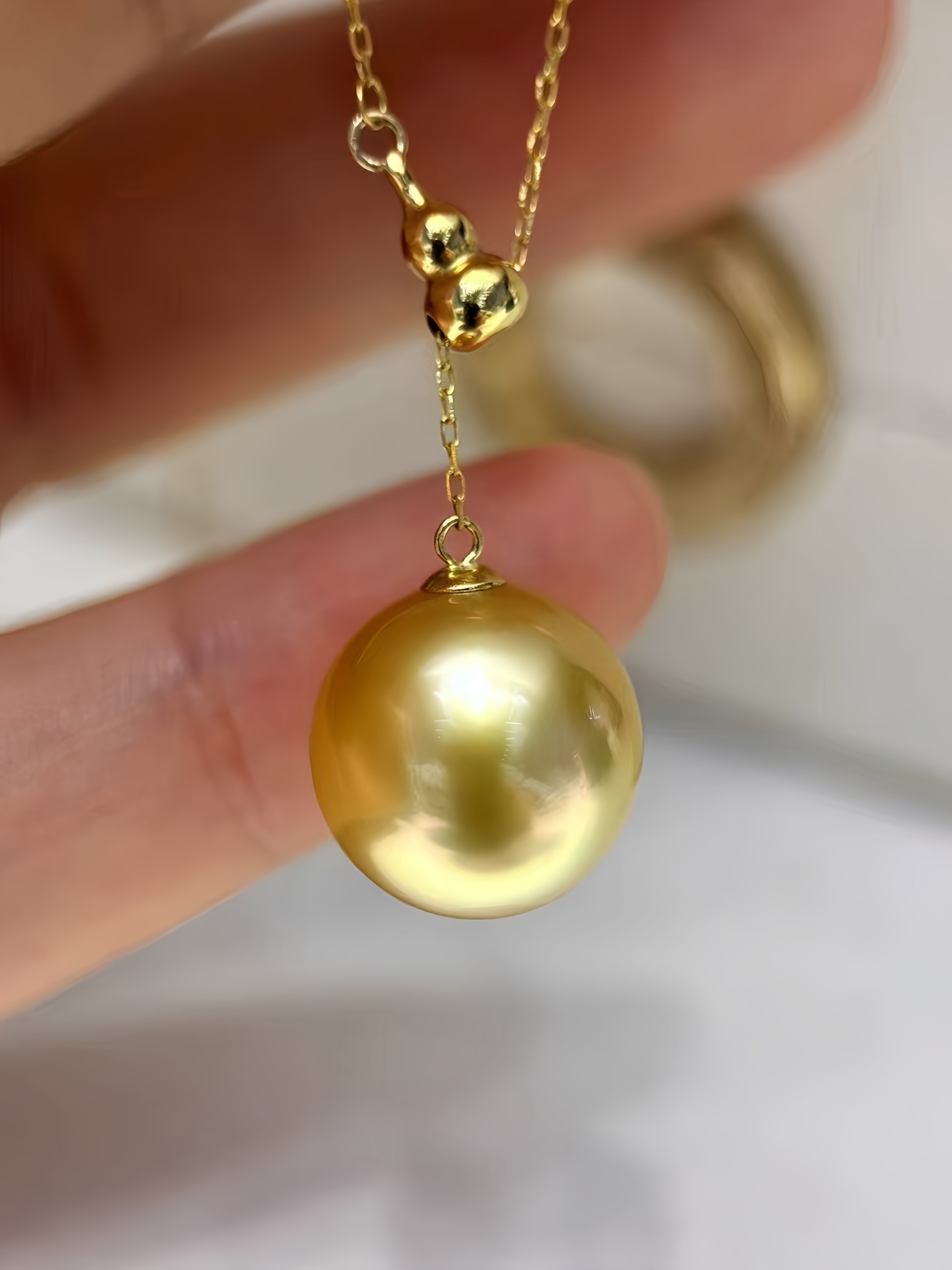 A gold pendant necklace featuring a large round freshwater pearl and two small gold beads on a delicate chain, held by a hand.