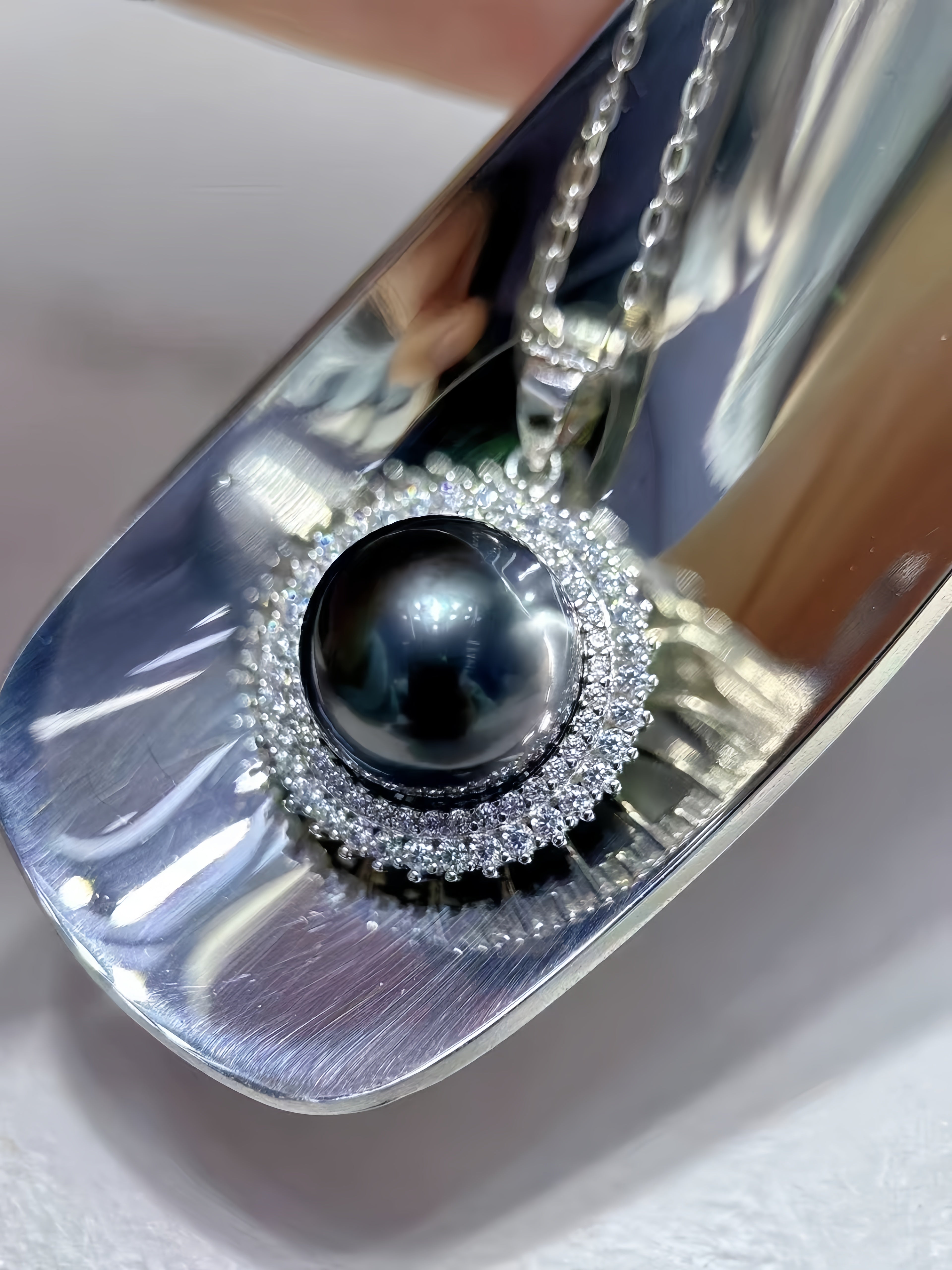 A close-up of a silver pendant necklace featuring a large black pearl surrounded by a halo of small clear stones, displayed on a reflective silver surface.
