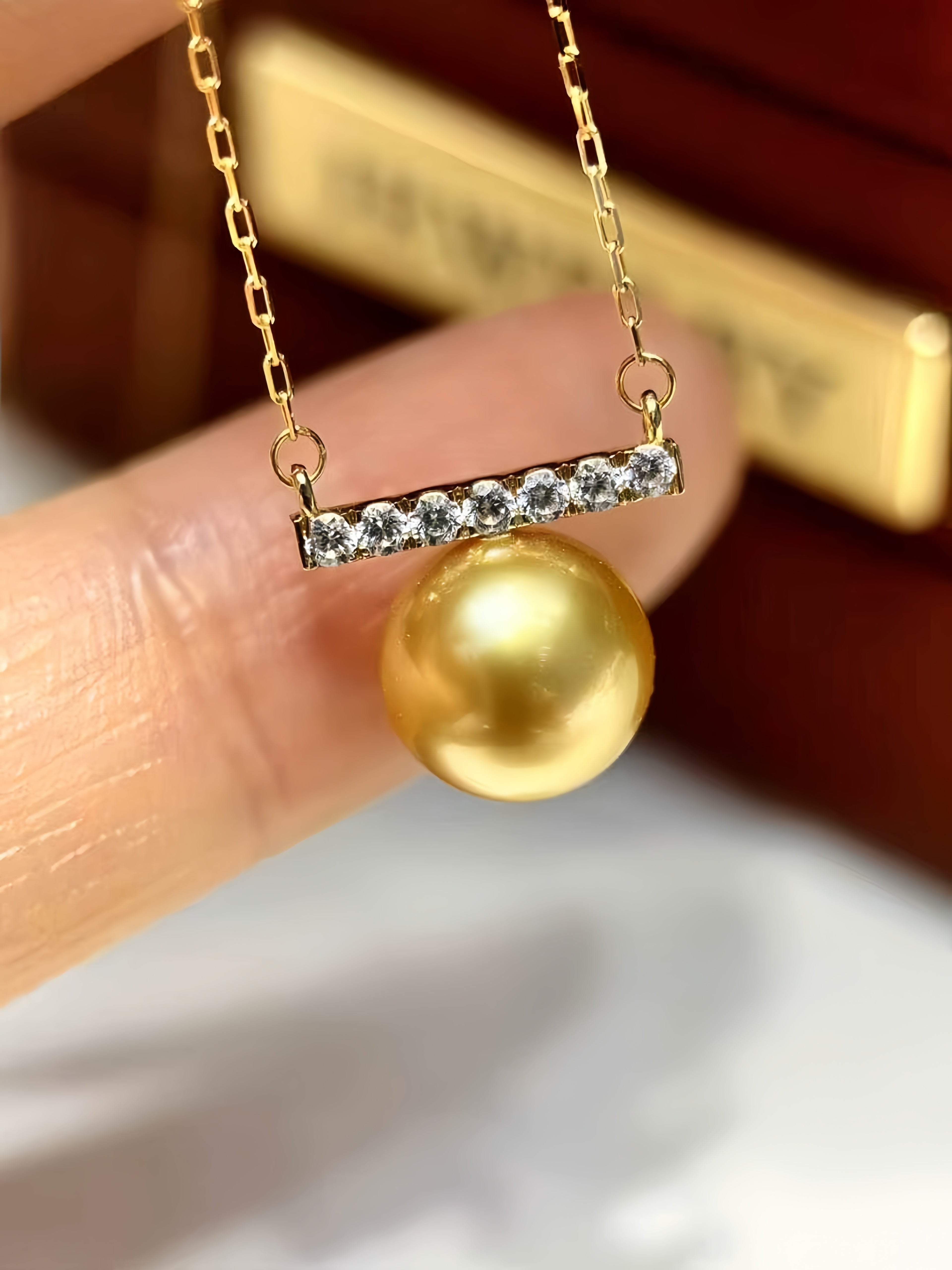 A gold pendant necklace featuring a bar set with small clear stones and a large round gold pearl, held between fingers against a blurred dark brown jewelry box background.