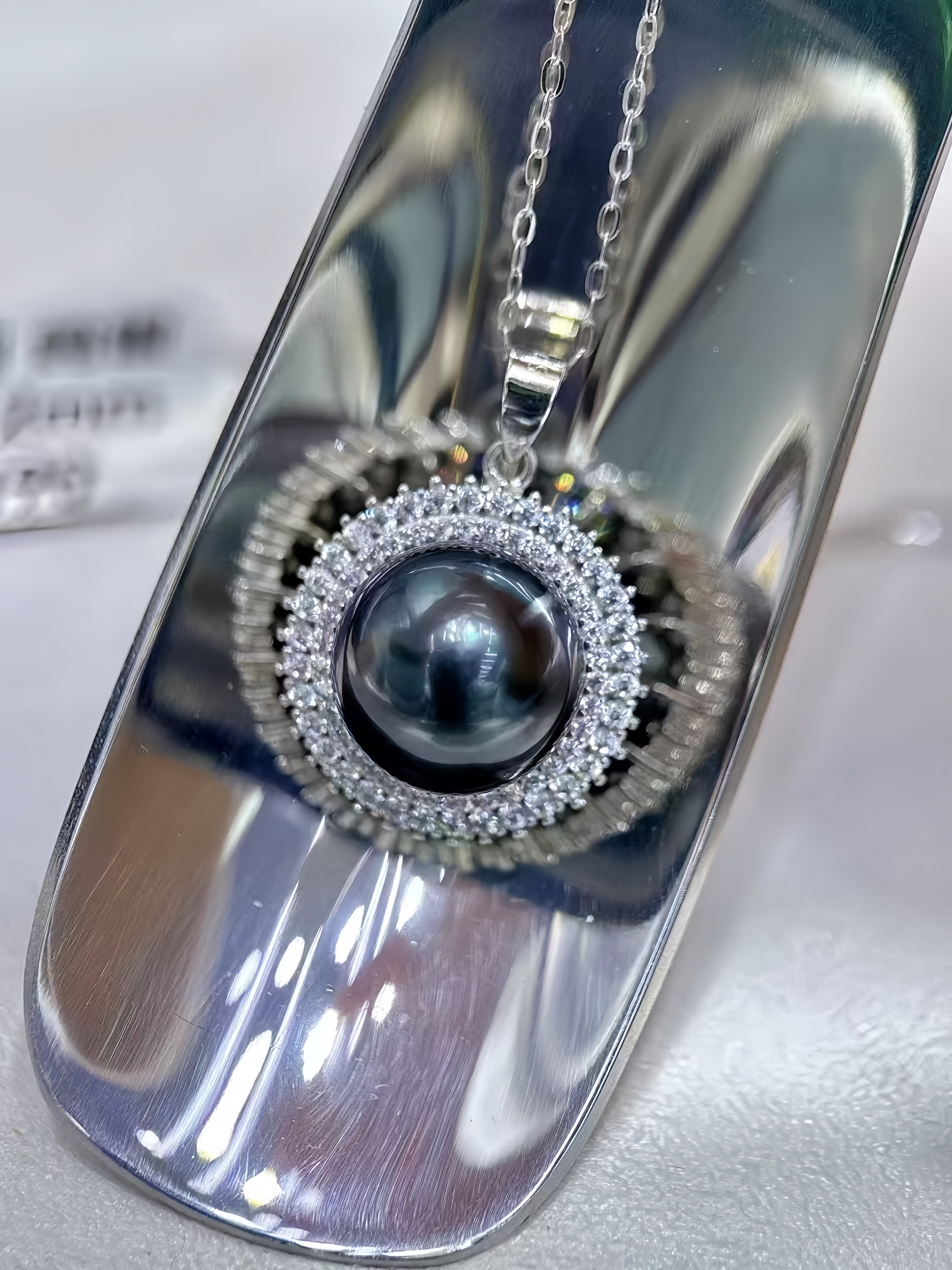 A silver pendant necklace featuring a dark gray pearl surrounded by a halo of small clear stones, displayed on a reflective silver stand.