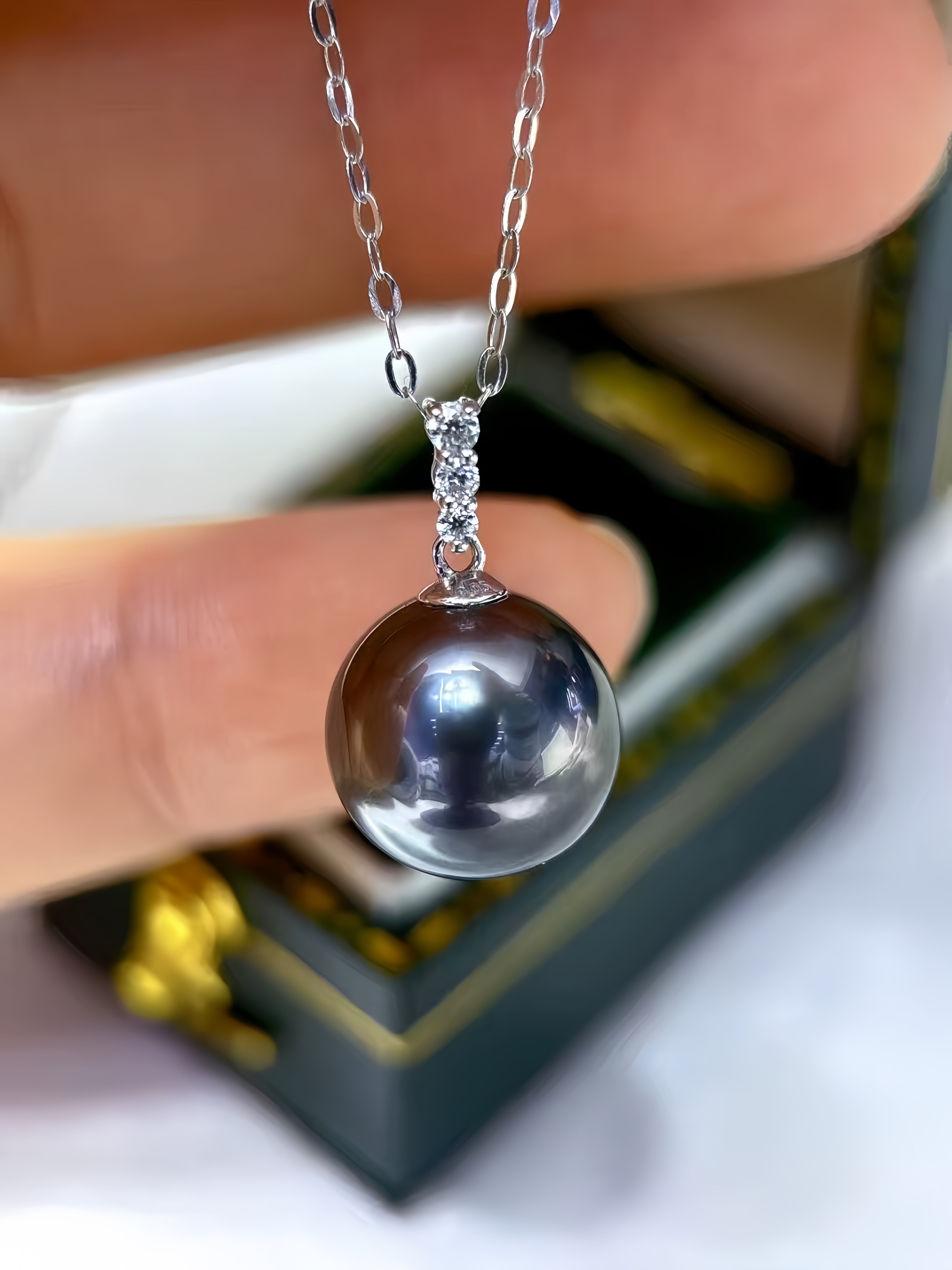 A close-up of a silver chain necklace with a round, dark gray pearl pendant held by fingers in front of a blurred jewelry box.