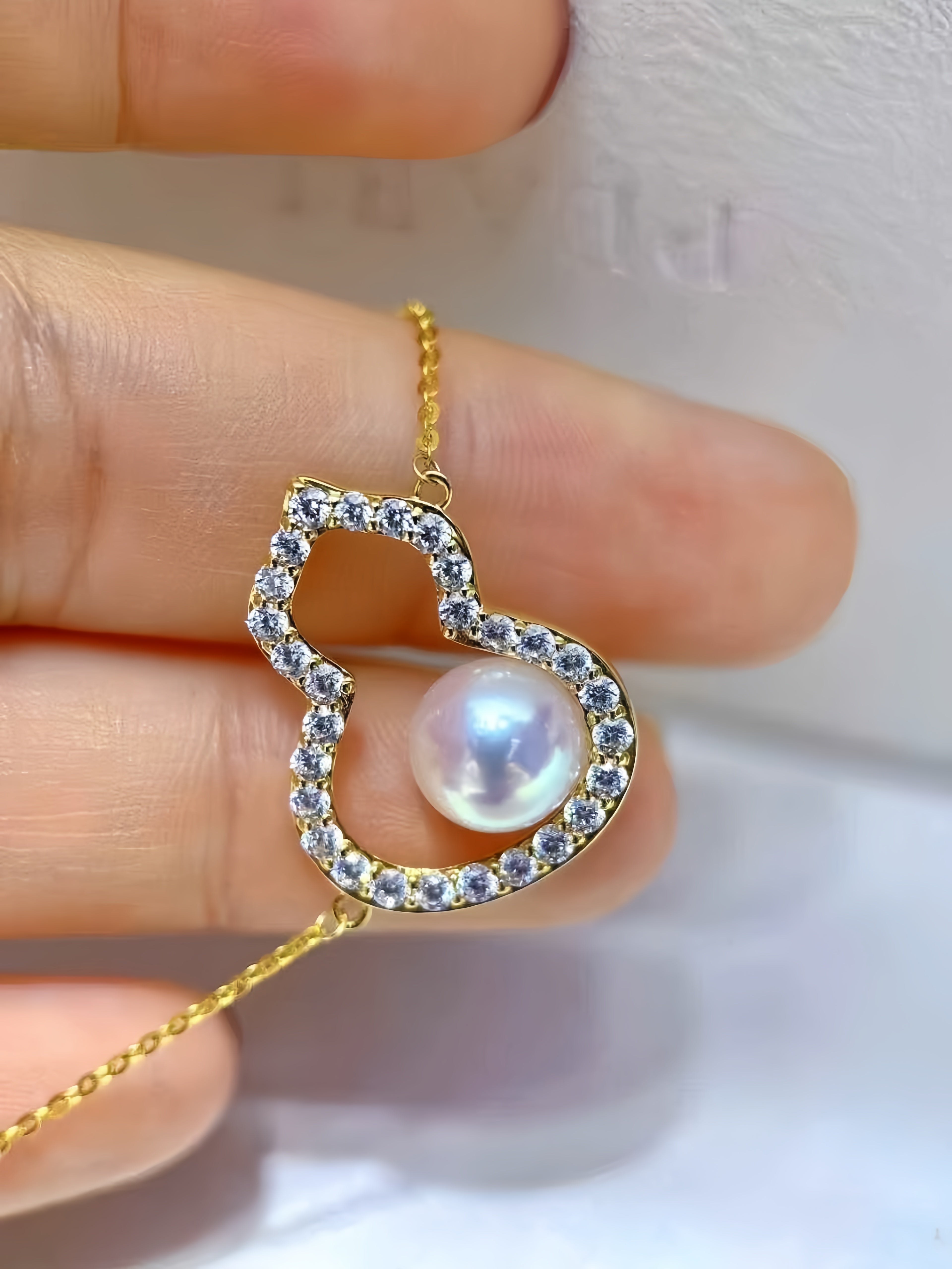 A gold heart-shaped pendant necklace with a central iridescent pearl, surrounded by small clear stones, held between fingers against a soft white background.